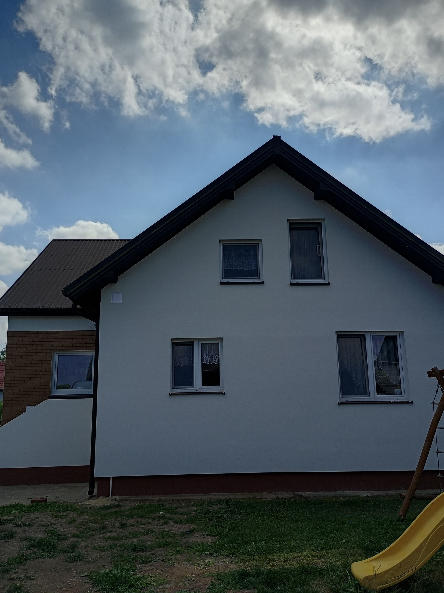 Biała elewacja domu jednorodzinnego z brązowym dachem w Miedzianej Górze. Widoczne okna z białymi firankami i fragment placu zabaw.