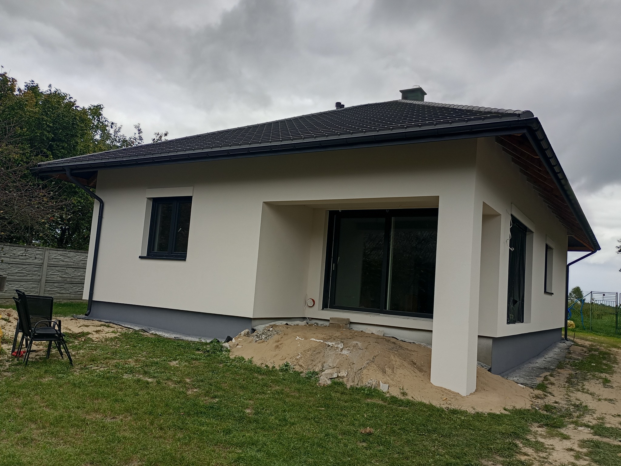 Jasny, jednopiętrowy dom z ciemną dachówką i szarą podmurówką, widziany z boku na tle pochmurnego nieba. Widoczne okna z czarnymi ramami i fragment ogrodu.