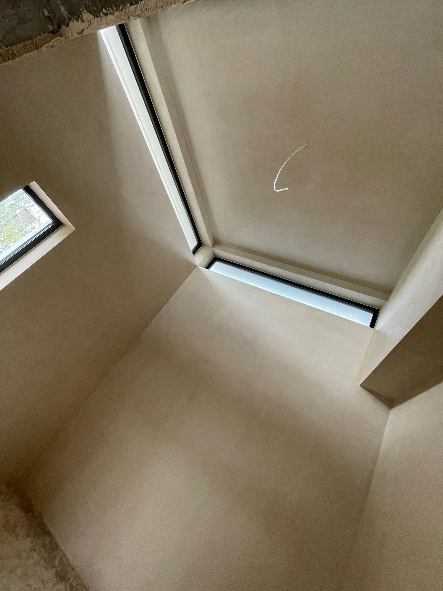 Wnętrze pokoju po nałożeniu jasnego, gładkiego tynku. Widoczne okna w czarnych ramach, osadzone w świeżo otynkowanych ścianach i suficie. Na suficie zwisa pojedynczy kabel.