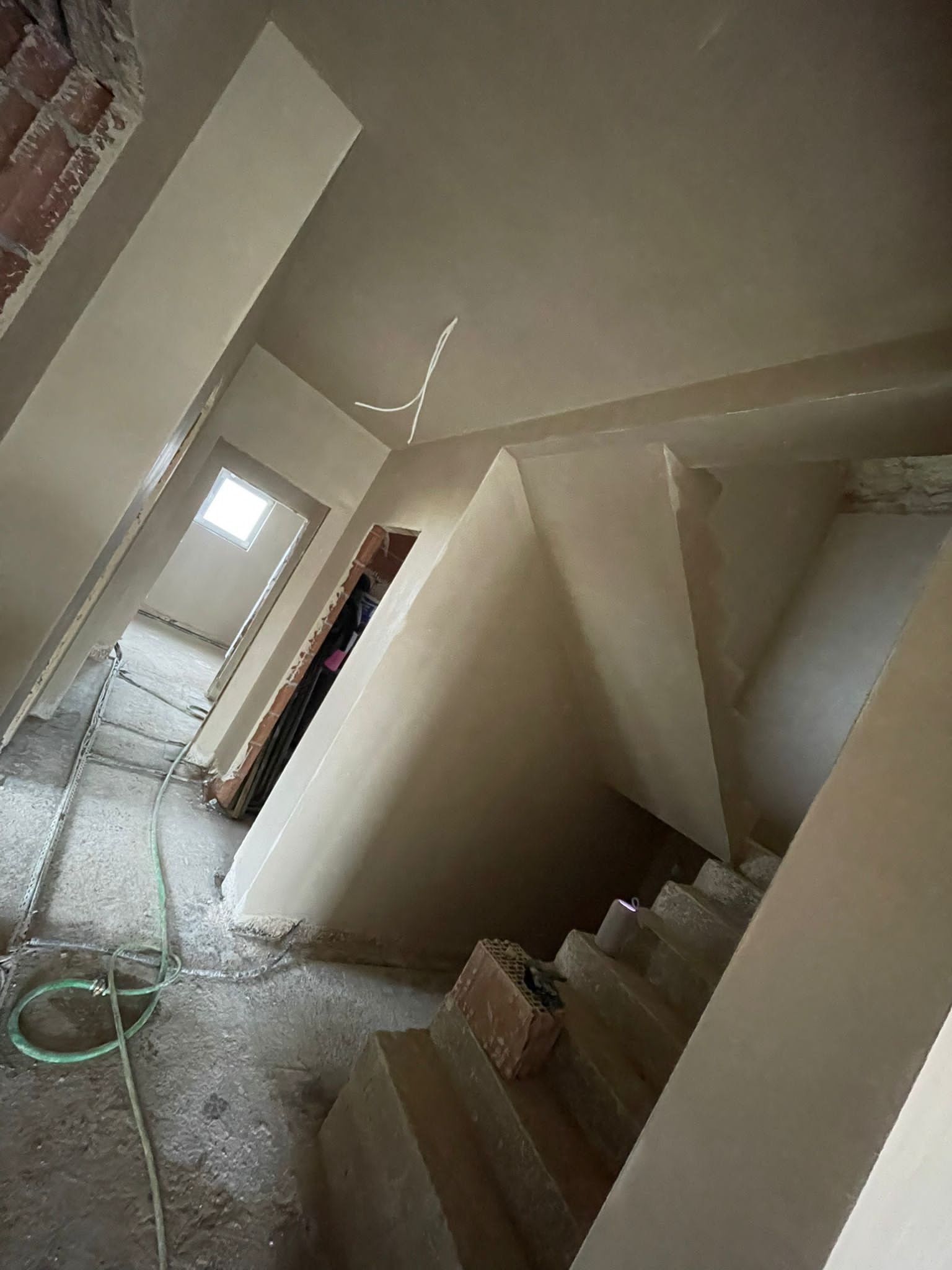 Wnętrze w trakcie prac wykończeniowych: świeżo otynkowane ściany i schody, widoczne fragmenty surowej cegły, zielony wąż na podłodze, perspektywa z dołu