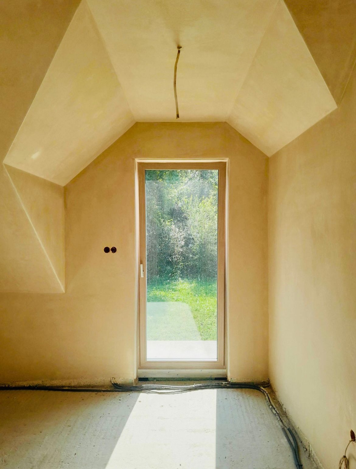 Świeżo otynkowane poddasze ze skośnym sufitem, widok na zewnątrz przez okno, widoczne kable elektryczne w ścianie i na podłodze, jasne wnętrze.