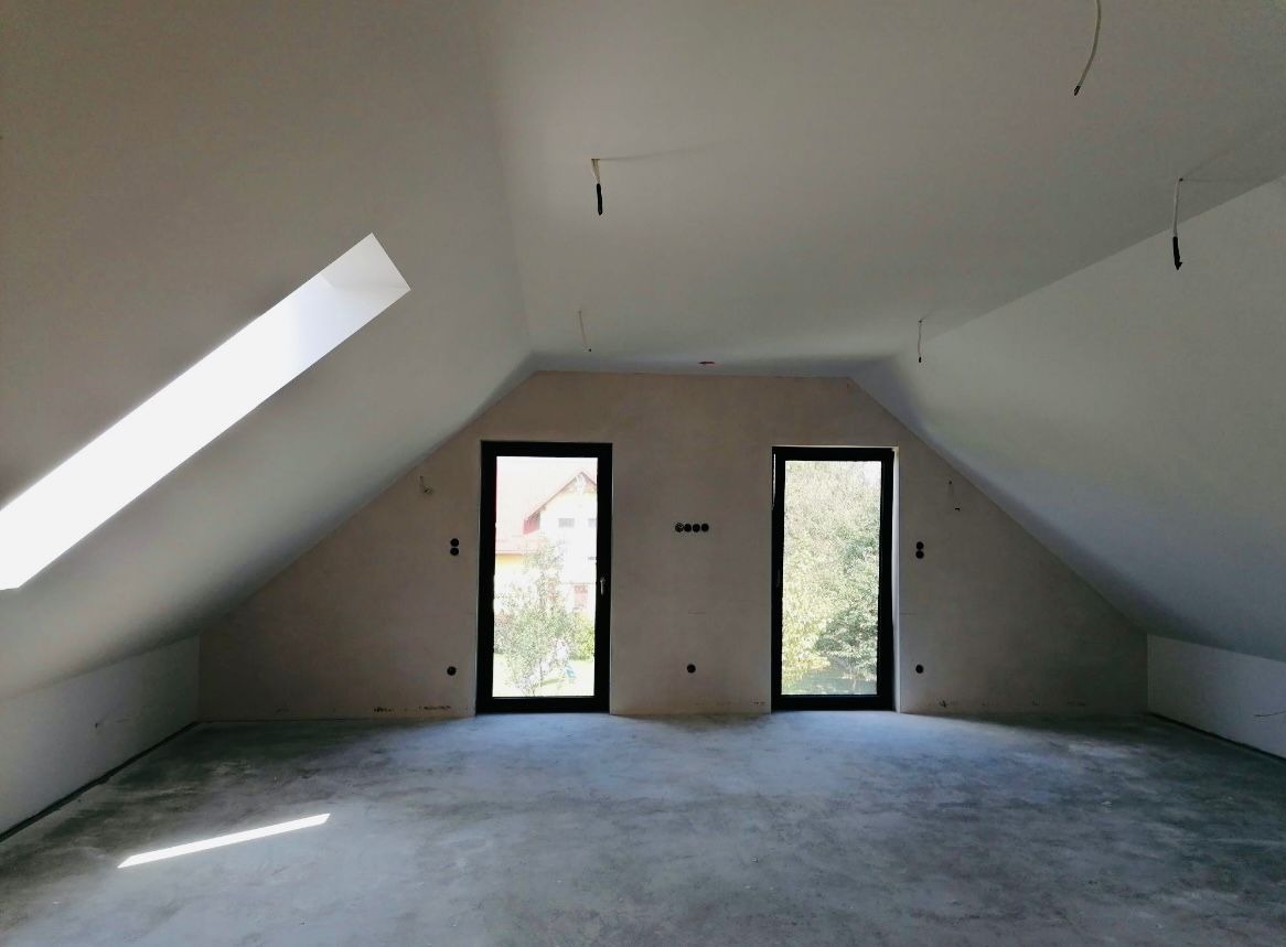 Poddasze w stanie surowym, ściany po nałożeniu gładzi, widoczne dwa okna i okno dachowe, przygotowane pod dalsze prace wykończeniowe.