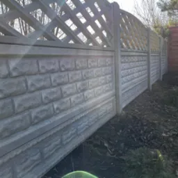 Długi, szary betonowy płot z dekoracyjnym wzorem kratki u góry i fakturą cegieł na dole, oświetlony słońcem, z widoczną ziemią pokrytą liśćmi i kulistymi lampami ogrodowymi.