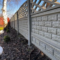 Michał Obarzanek Clean Brothers - Długi, betonowy płot panelowy z ażurową górą, imitujący cegłę, oddzielający posesję, widoczny z perspektywy ziemi, z brzozą i elementami ogrodu w tle.