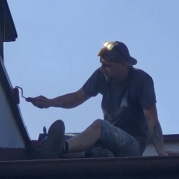 Michał Obarzanek Clean Brothers - Mężczyzna z czołówką maluje dach za pomocą wałka, siedząc na jego krawędzi. Widok z dołu, na tle jasnego nieba.