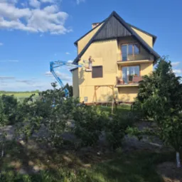 Pracownik na podnośniku czyści elewację dwupiętrowego domu z balkonami. Widok z ogrodu z krzewami, w tle pola i błękitne niebo z chmurami.