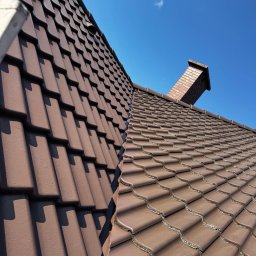 Michał Obarzanek Clean Brothers - Ciemnobrązowy dach z dachówki ceramicznej, widok z dołu na tle błękitnego nieba z kominem. Układanka dachowa z wyraźną fakturą i cieniem.