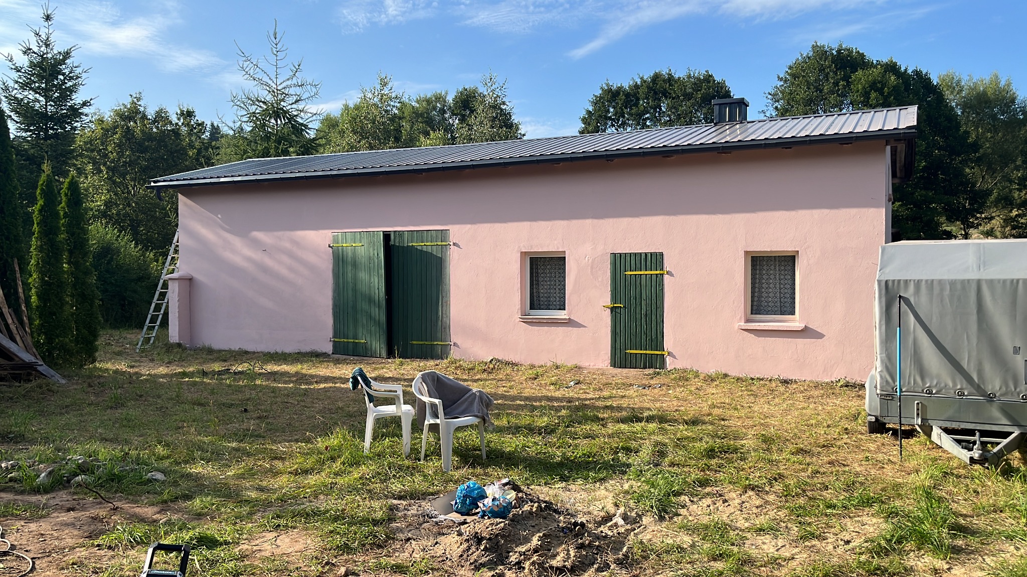 Różowa elewacja budynku gospodarczego z zielonymi okiennicami i szarym dachem. Na trawniku leżą rzeczy, a z boku stoi przyczepa.