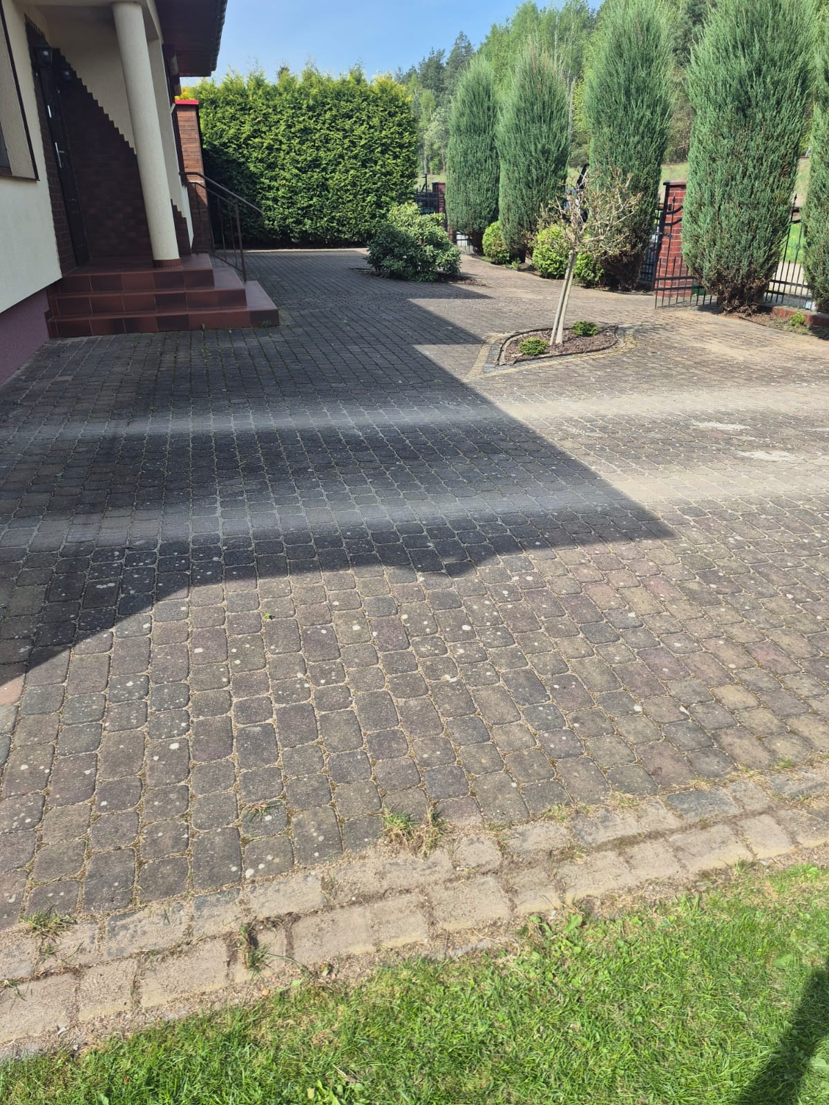 Szara kostka brukowa na podjeździe, z widocznym cieniem, obok domu z zielenią. Część kostki jest bardziej zabrudzona, co sugeruje potrzebę czyszczenia.