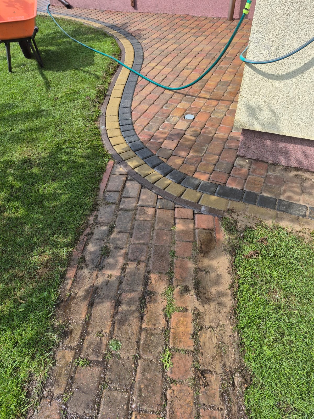 Częściowo umyta kostka brukowa z obrzeżem w odcieniach żółci i szarości, widoczny wąż ogrodowy i fragment trawnika. Na pierwszym planie widoczny brudny fragment kostki.
