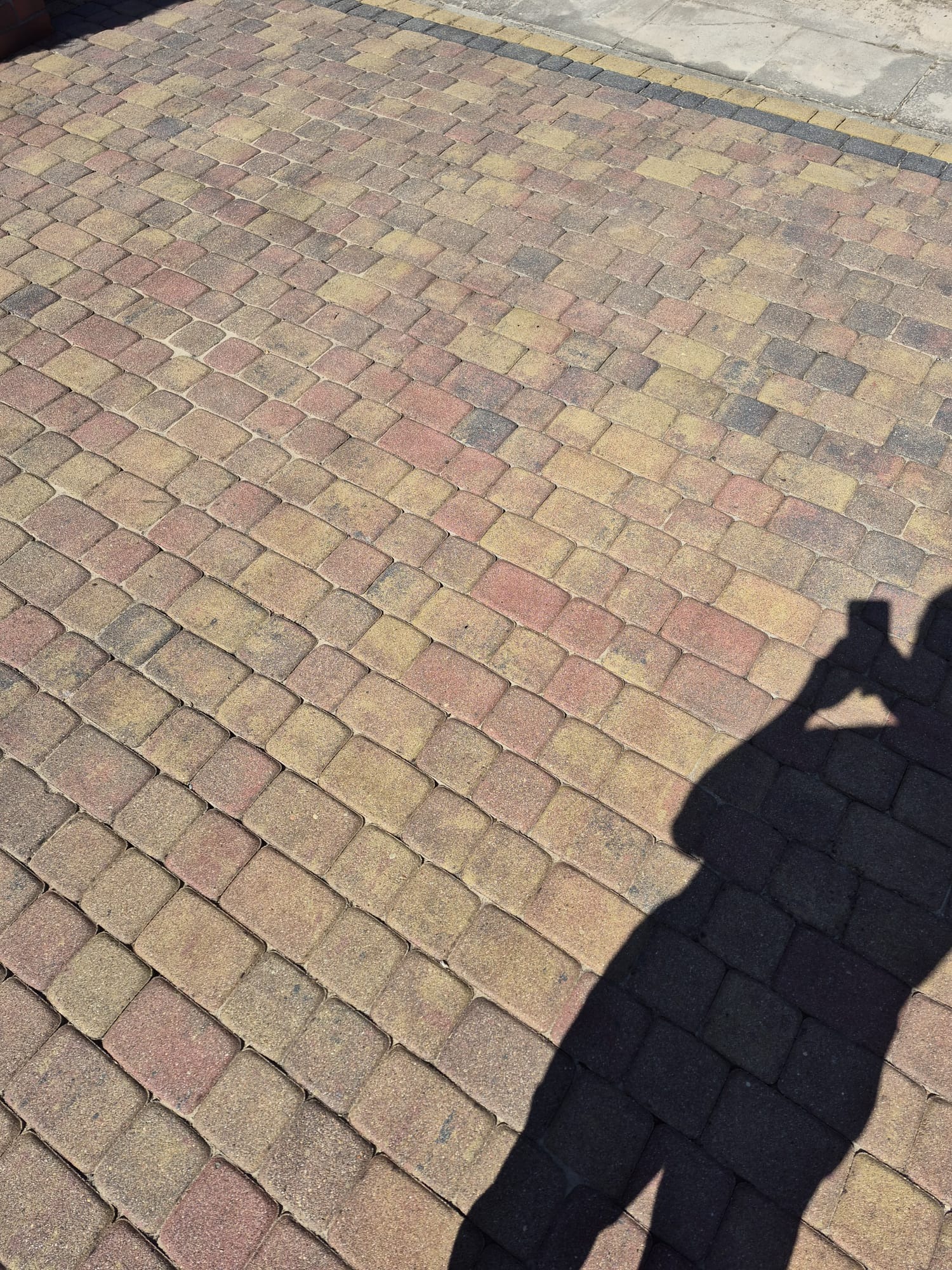 Ułożona kostka brukowa w odcieniach żółci i czerwieni, widziana pod kątem, z cieniem osoby robiącej zdjęcie w prawym dolnym rogu. Widoczne krawężniki.