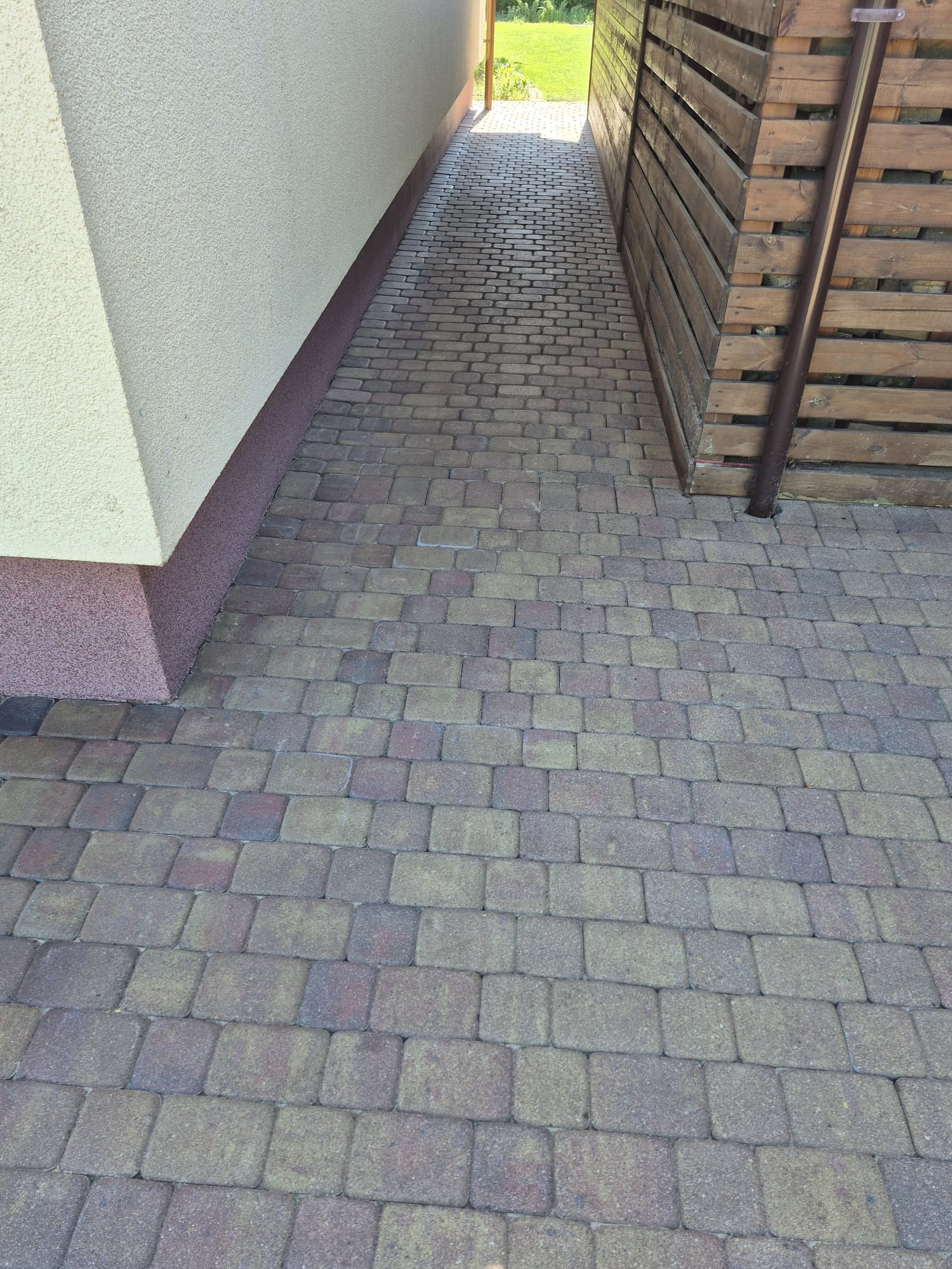 Ułożona kostka brukowa w wąskim przejściu między budynkiem z beżową elewacją a drewnianym płotem. Widoczny fragment rynny. Kostka w odcieniach szarości i brązu.