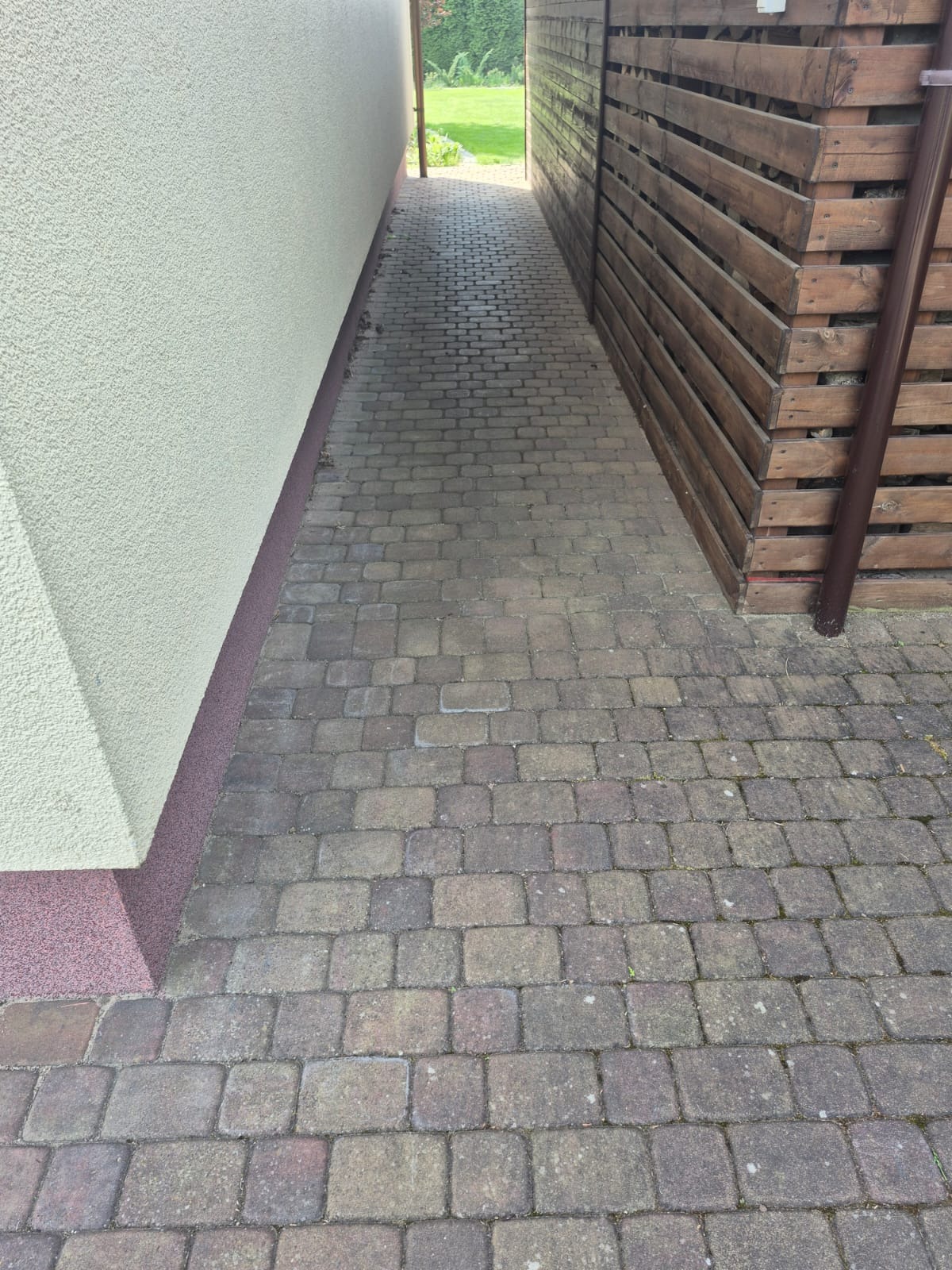 Ułożona kostka brukowa tworząca ścieżkę między budynkiem z jasną elewacją a drewnianym ogrodzeniem. Widoczny fragment rynny. Perspektywa z dołu w stronę zieleni.