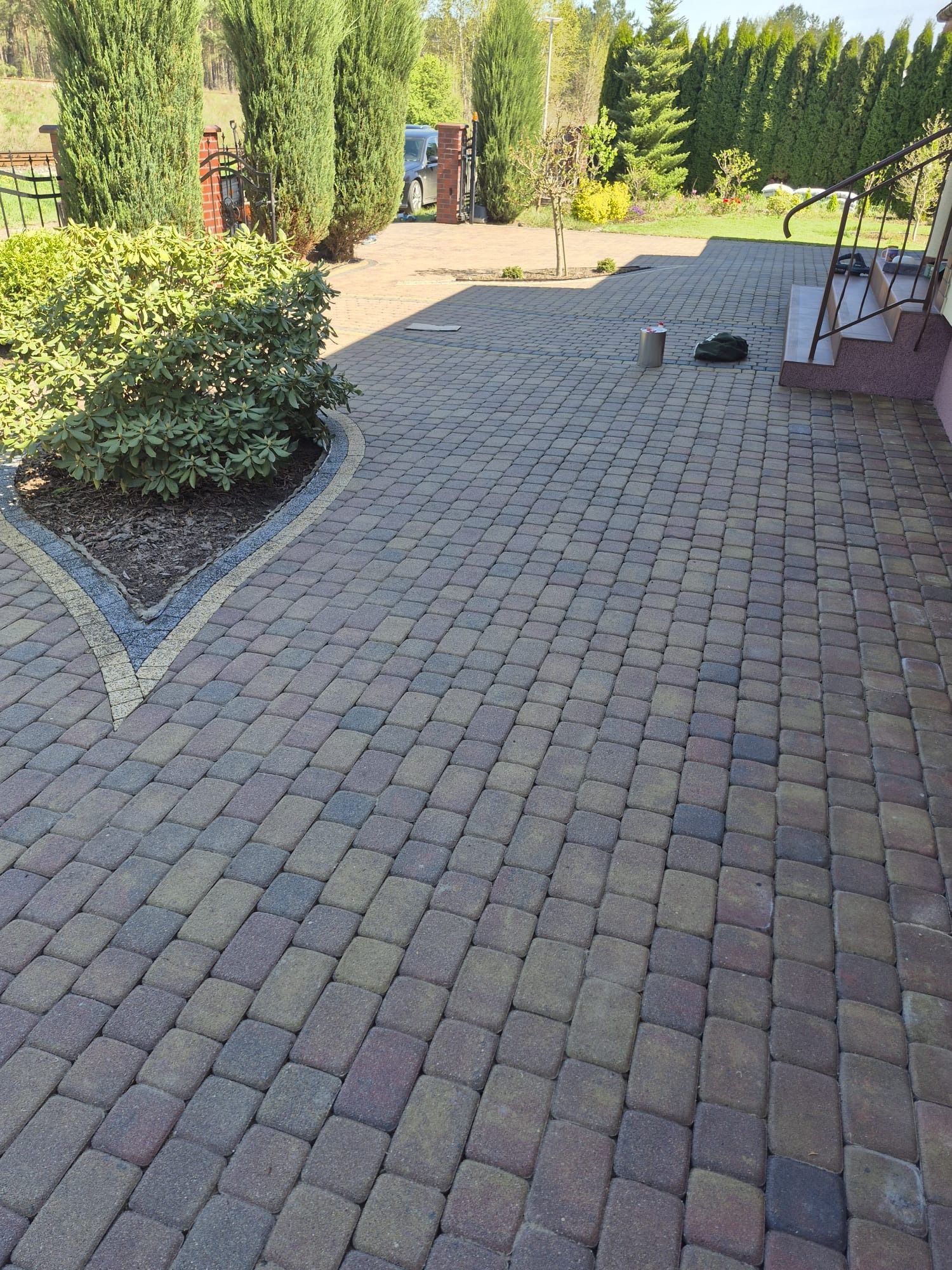 Ułożona kostka brukowa na podjeździe, z widoczną roślinnością i schodami wejściowymi do domu. Na kostce stoi puszka i torba.