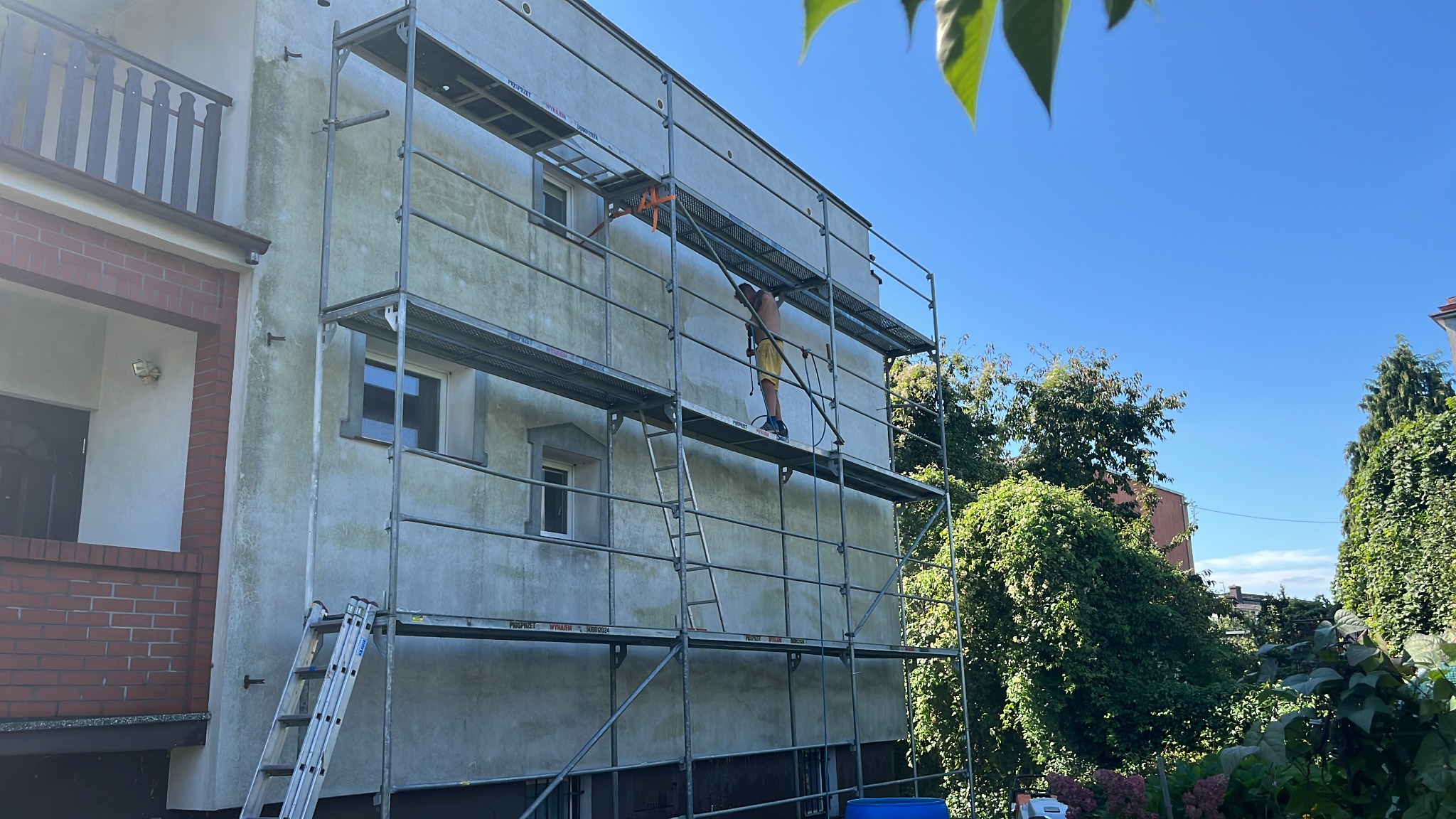 Pracownik na rusztowaniu czyści elewację budynku z użyciem myjki ciśnieniowej. Widoczne zabrudzenia na ścianie przed czyszczeniem.