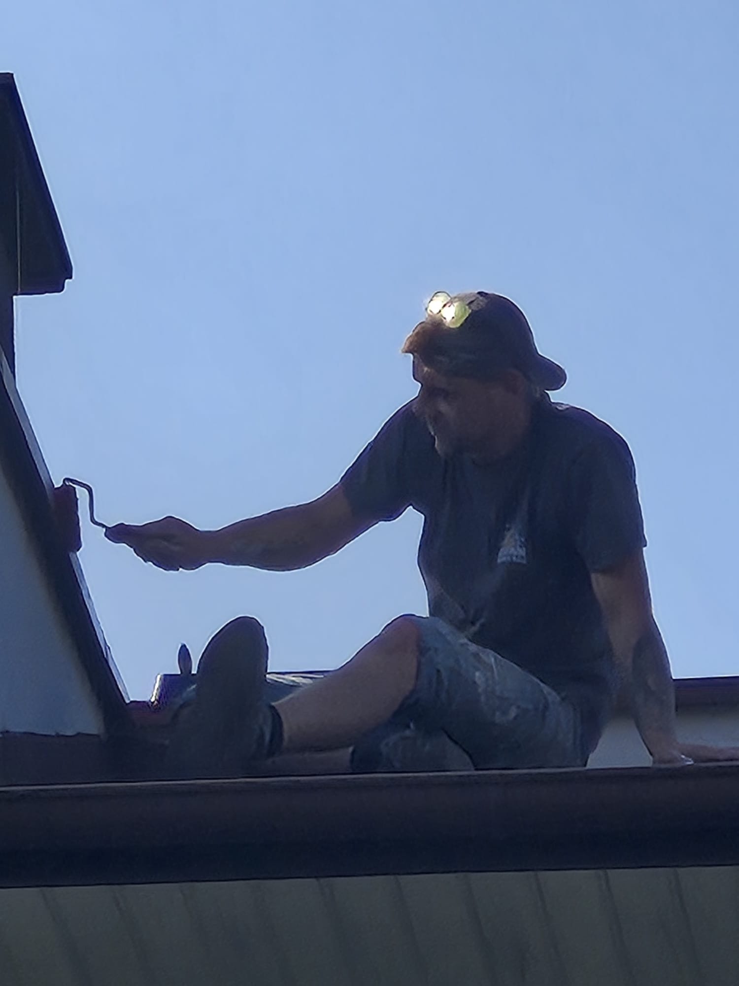 Mężczyzna z czołówką maluje dach za pomocą wałka, siedząc na jego krawędzi. Widok z dołu, na tle jasnego nieba.