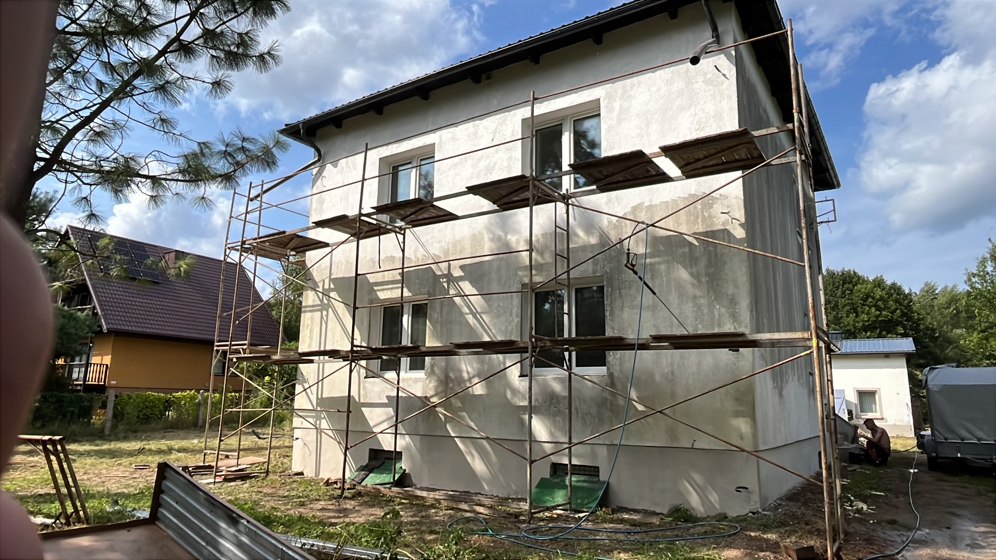 Dom w Debrznie w trakcie prac elewacyjnych. Widoczne rusztowania i surowa, jasna elewacja. W tle budynek z panelami słonecznymi i błękitne niebo.