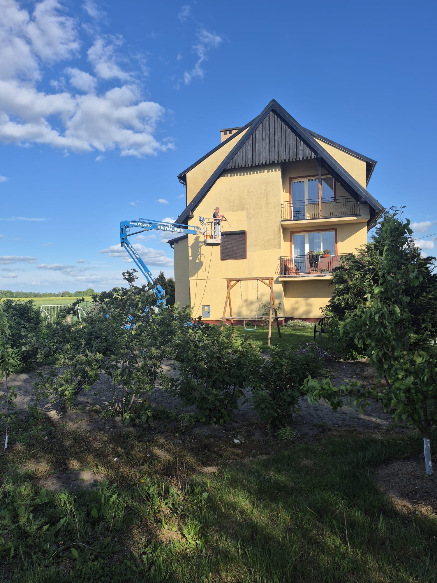 Pracownik na podnośniku czyści elewację dwupiętrowego domu z balkonami. Widok z ogrodu z krzewami, w tle pola i błękitne niebo z chmurami.