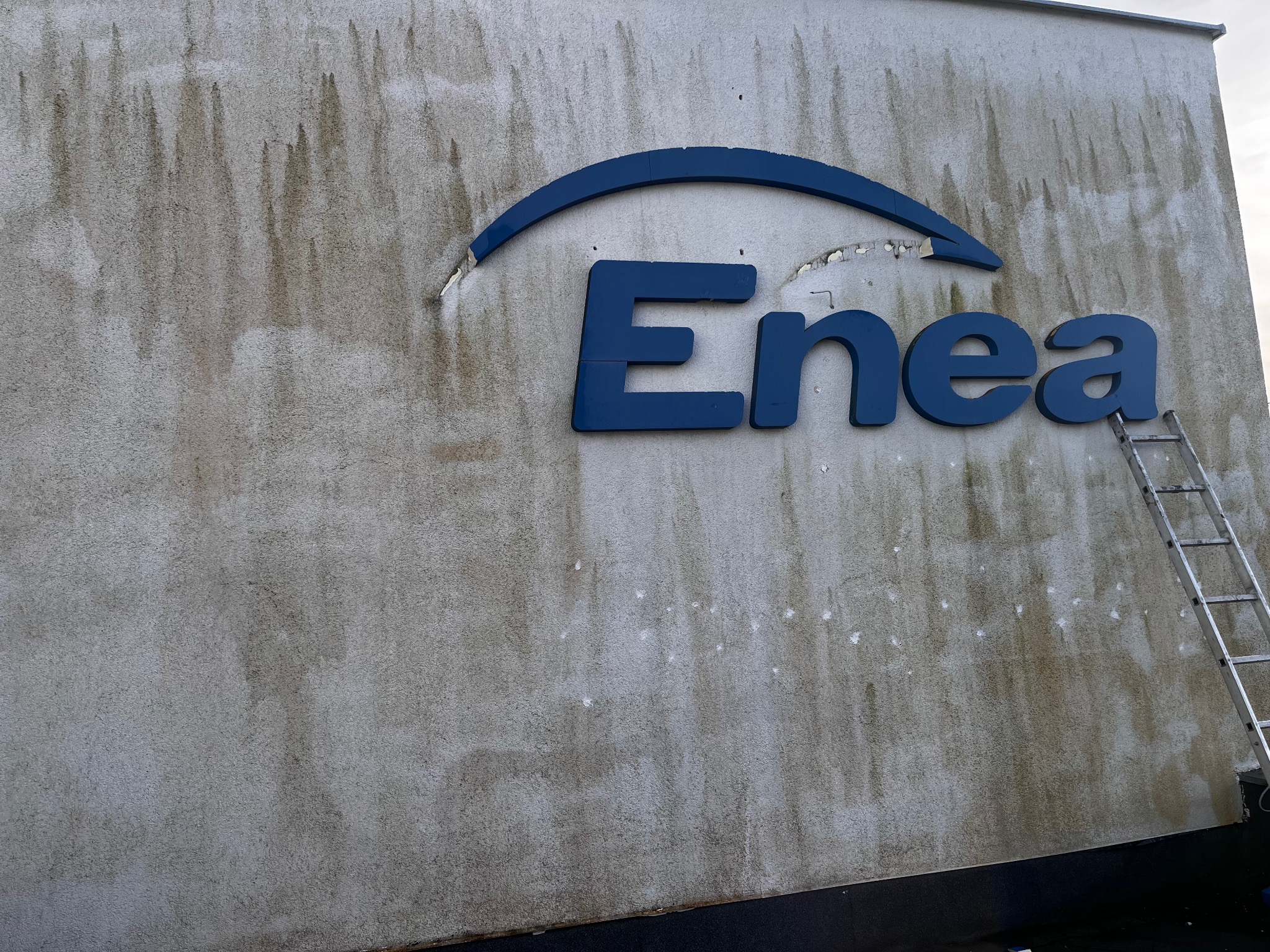 Brudna elewacja budynku z logo firmy Enea, częściowo uszkodzonym. Widoczna drabina oparta o ścianę. Ślady zacieków i zabrudzeń na elewacji.