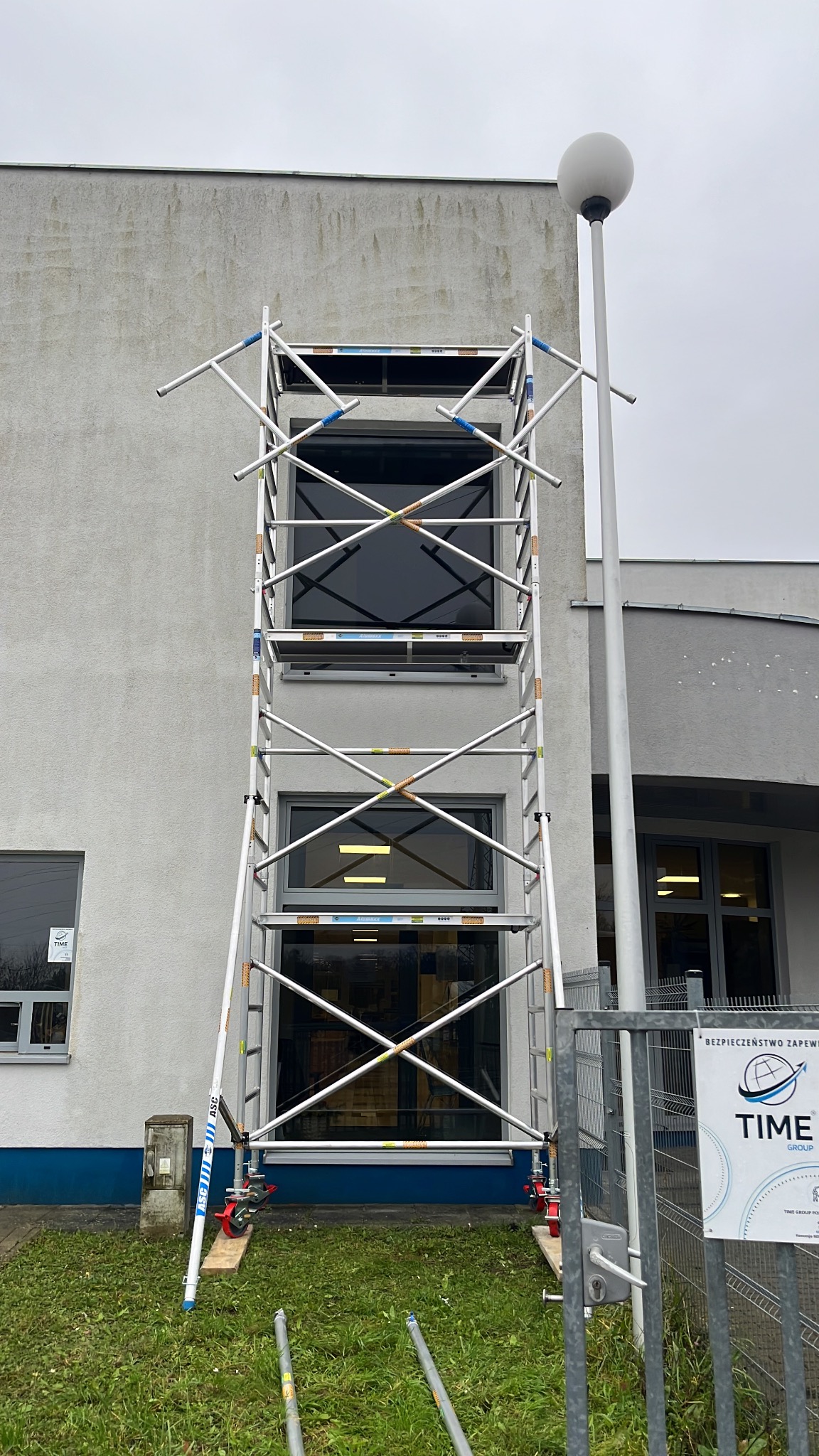 Aluminiowe rusztowanie ustawione przed budynkiem z dwoma oknami. Widoczne elementy rusztowania, fragment elewacji i lampa uliczna. Ujęcie z dołu, pochmurne niebo.