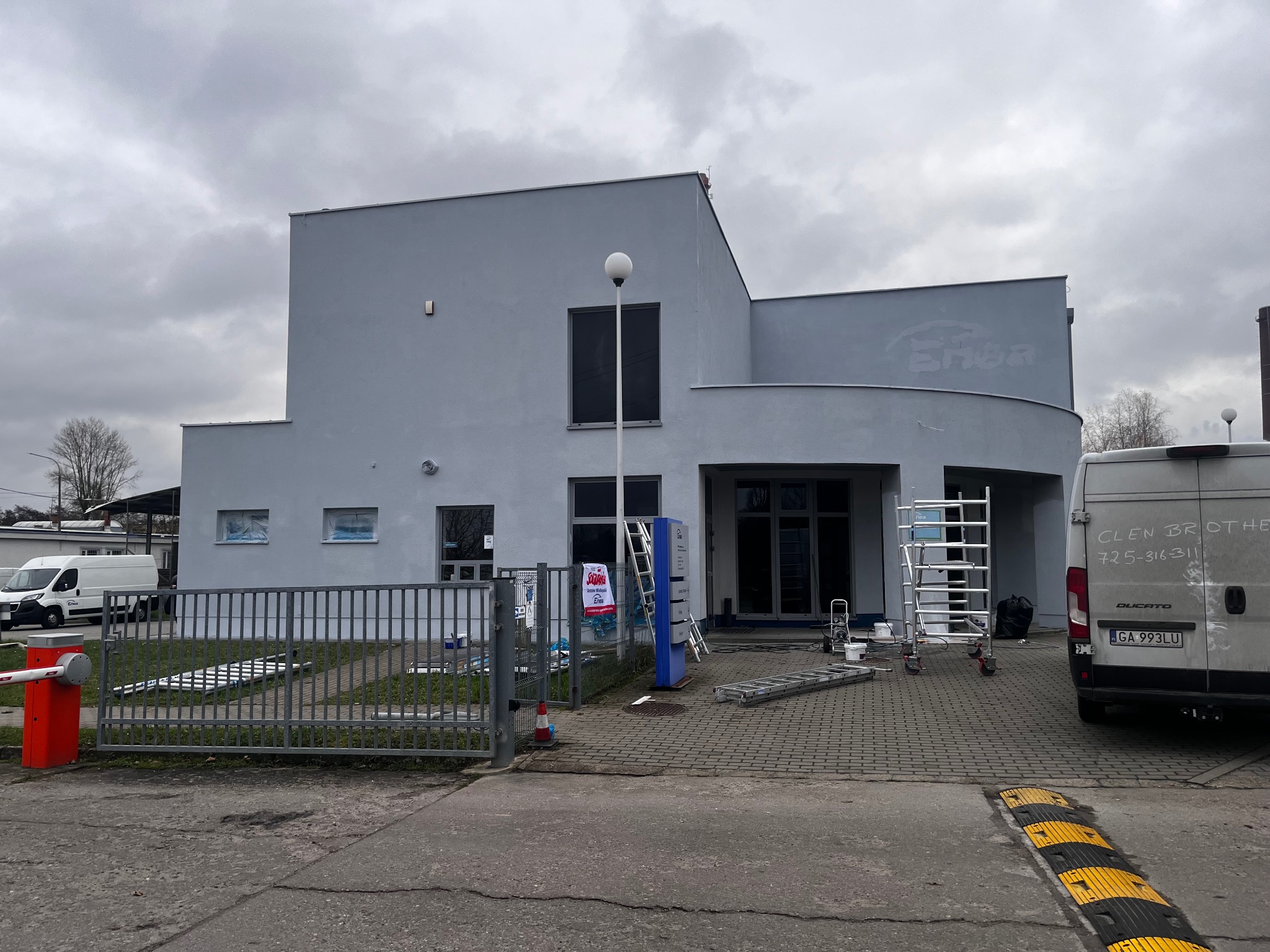 Budynek po malowaniu elewacji na szaro. Widoczne rusztowania, drabiny i samochód dostawczy. Okna zabezpieczone folią. Pochmurne niebo w tle.