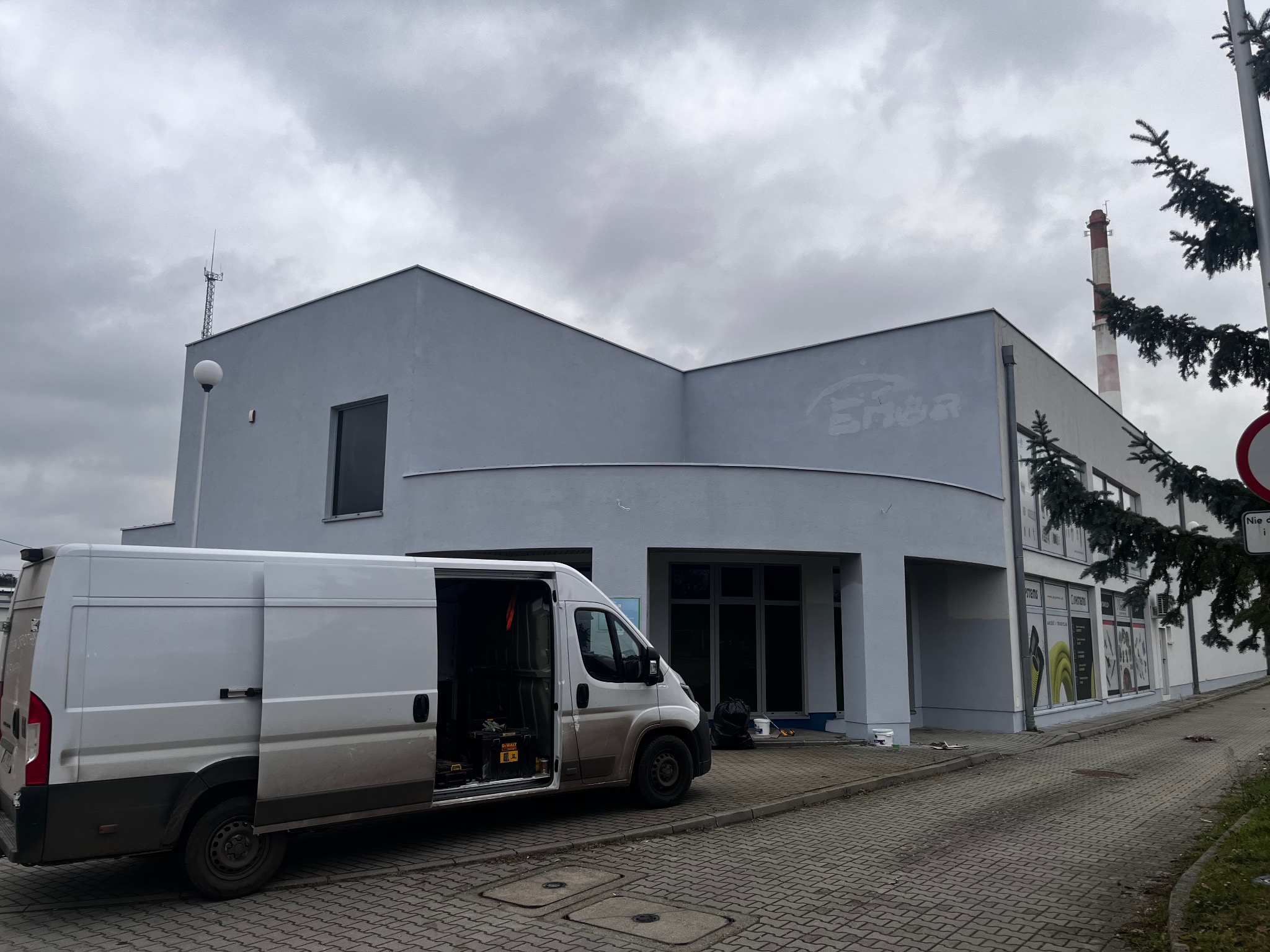 Szary budynek z otwartym busem serwisowym na chodniku. Widoczne ślady po usuniętym napisie na elewacji. W tle komin i pochmurne niebo.