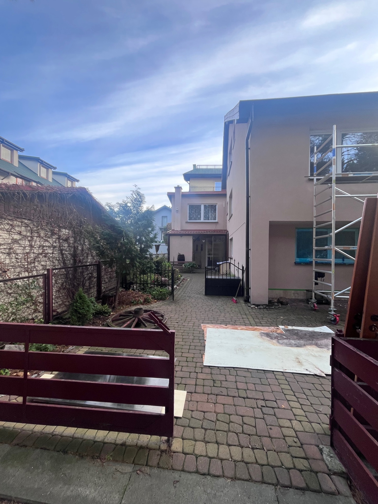 Jasny budynek z rusztowaniem w trakcie prac elewacyjnych, widok z brukowanego podwórza z elementami dekoracyjnymi i zielenią. Częściowo zasłonięte ogrodzenie.