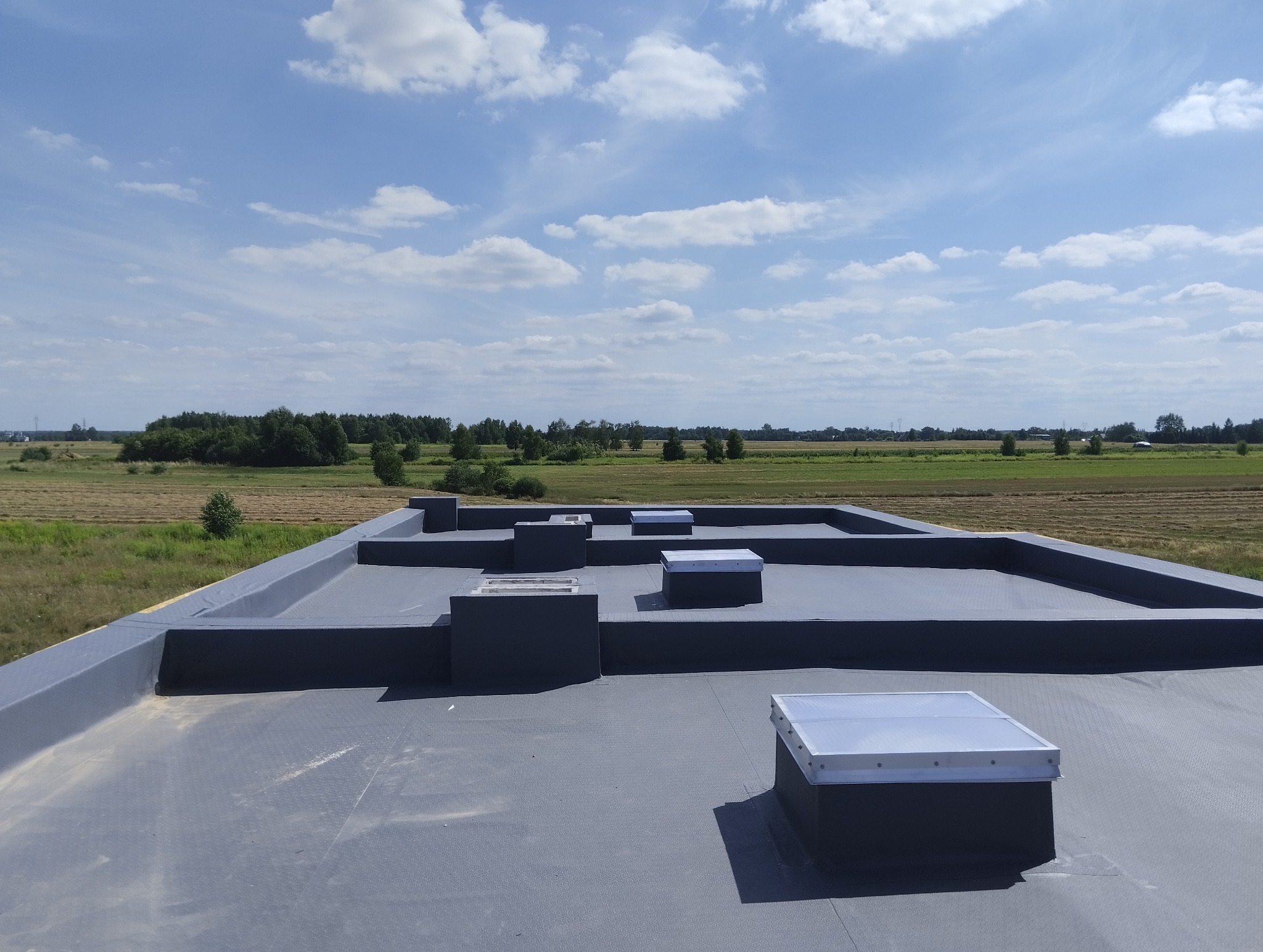 Płaski dach pokryty szarą membraną z świetlikami dachowymi i obróbkami blacharskimi, widok na pola i błękitne niebo z chmurami.