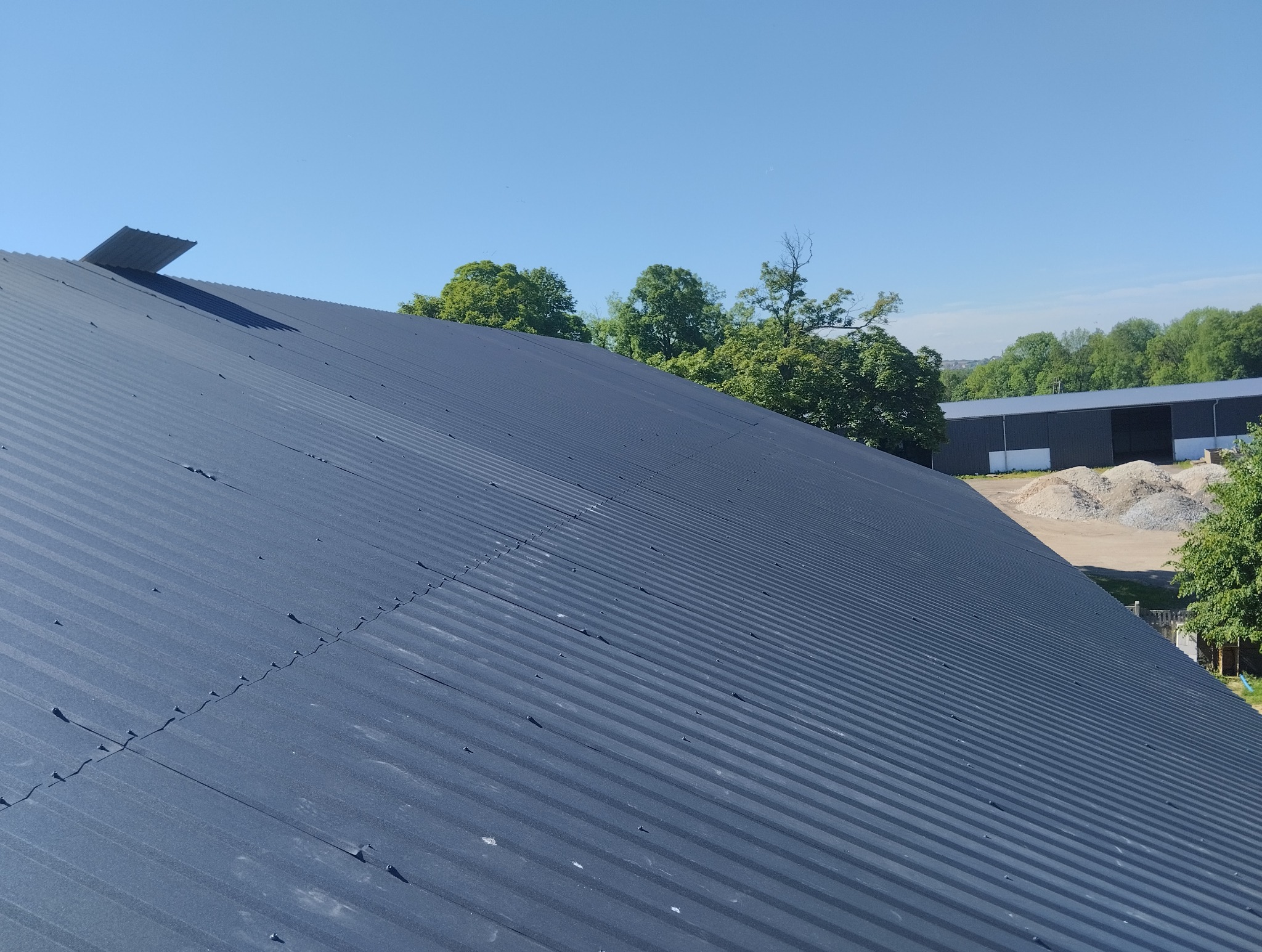 Nowy, ciemnoszary dach z blachy trapezowej, widok z góry, w tle drzewa i budynek magazynowy.