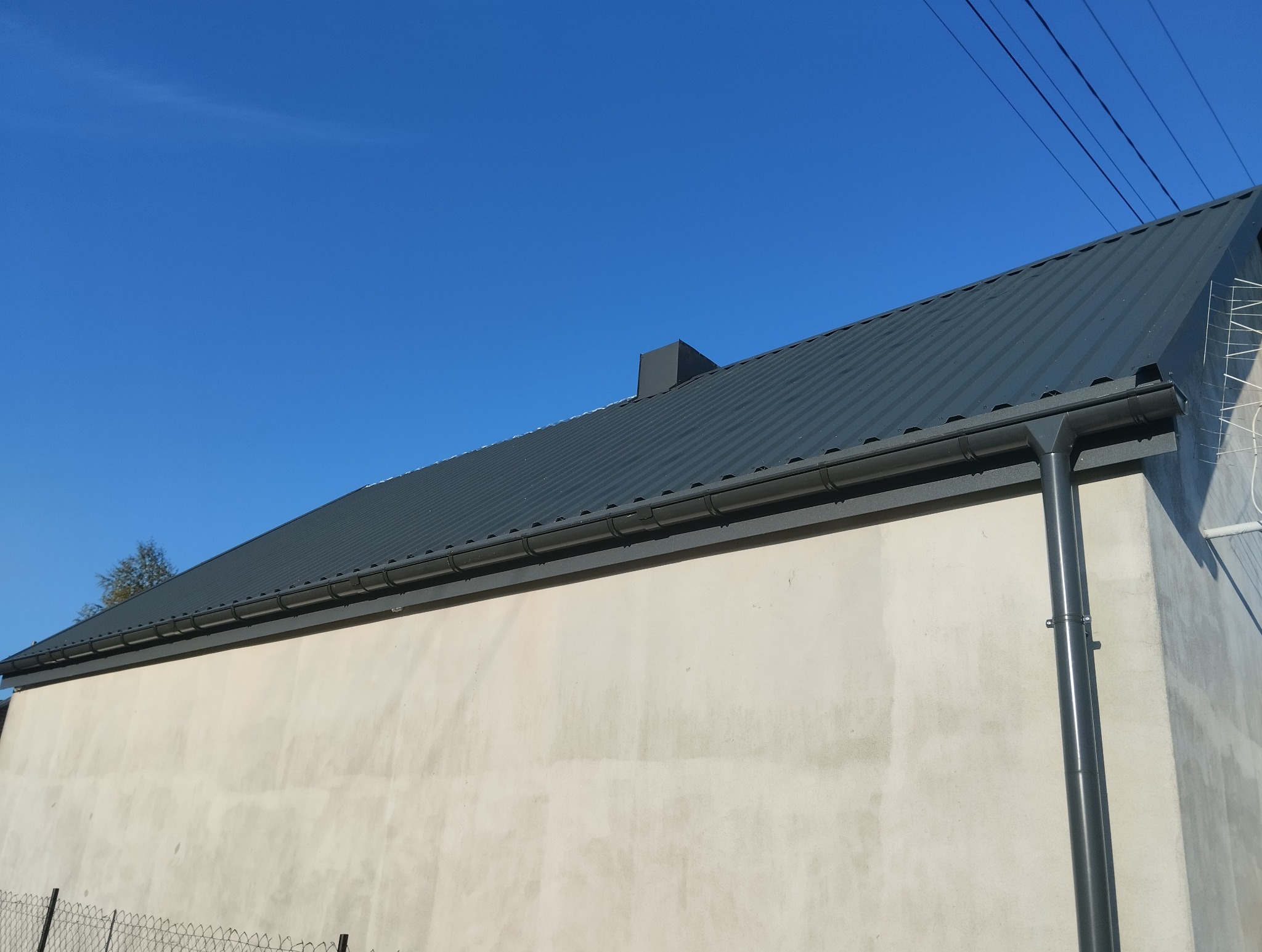 Widok z dołu na szary dach z blachy trapezowej, szare rynny i komin na tle błękitnego nieba, widoczna ściana budynku z anteną.