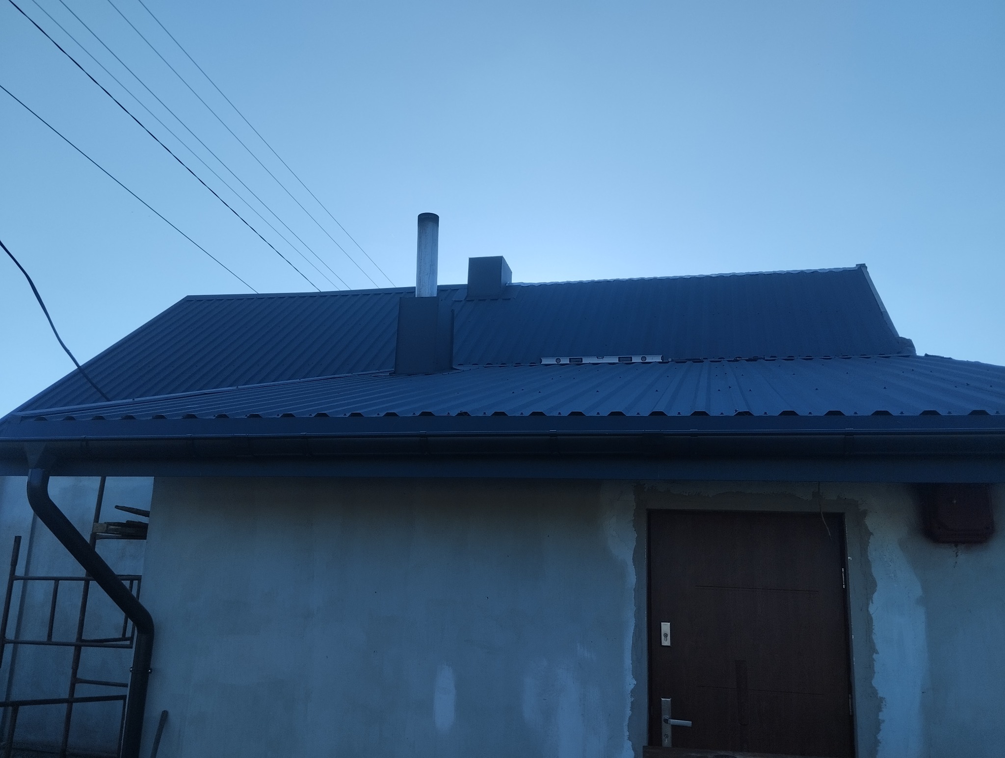 Nowy, ciemny dach z blachy trapezowej z kominem i rynnami, widoczna drabina oparta o ścianę budynku.