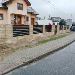 AMTRANS ANTONI OGIERMAN - Nowoczesne ogrodzenie z betonowych bloczków i poziomych, czarnych przęseł metalowych, widok na dom z beżową elewacją, pochmurny dzień.
