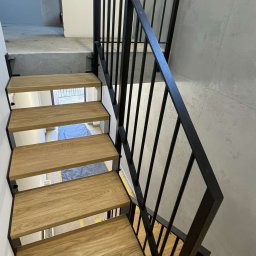 AMTRANS ANTONI OGIERMAN - Nowoczesne schody z drewnianymi stopniami i czarną, metalową balustradą, widok z dołu, fragment podłogi zabezpieczony folią malarską.