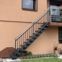 AMTRANS ANTONI OGIERMAN - Zewnętrzne schody metalowe z poręczą ze stali nierdzewnej prowadzące na balkon, przymocowane do elewacji budynku z dekoracyjnym oknem i roślinami w donicach.