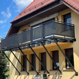 AMTRANS ANTONI OGIERMAN - Montaż balkonu z czarną, metalową balustradą na żółtej elewacji budynku, widoczne ukośne wsporniki i dwóch monterów na podnośniku.