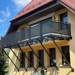 Montaż balkonu z czarną, metalową balustradą na żółtej elewacji budynku, widoczne ukośne wsporniki i dwóch monterów na podnośniku.