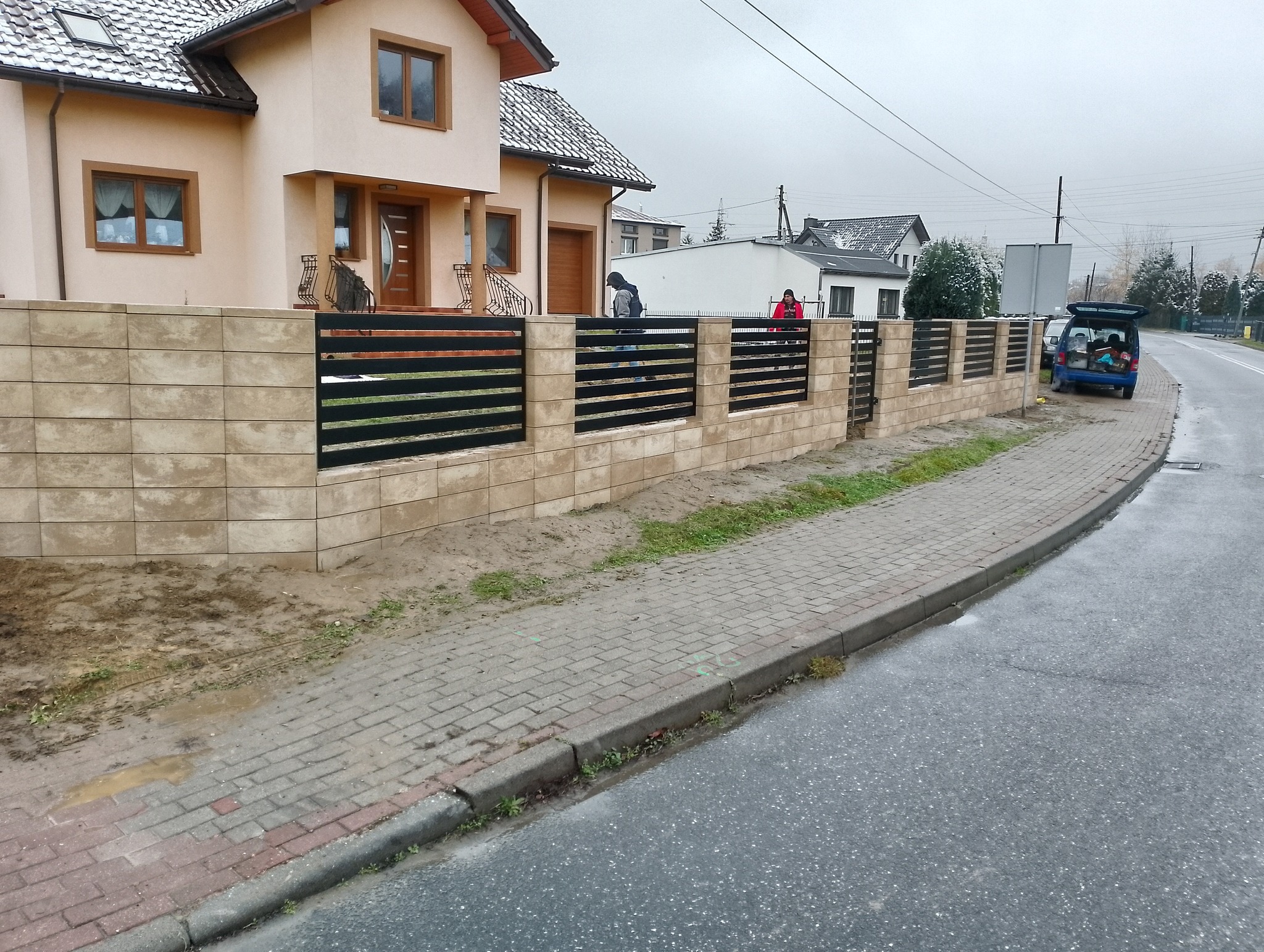 Nowoczesne ogrodzenie z betonowych bloczków i poziomych, czarnych przęseł metalowych, widok na dom z beżową elewacją, pochmurny dzień.