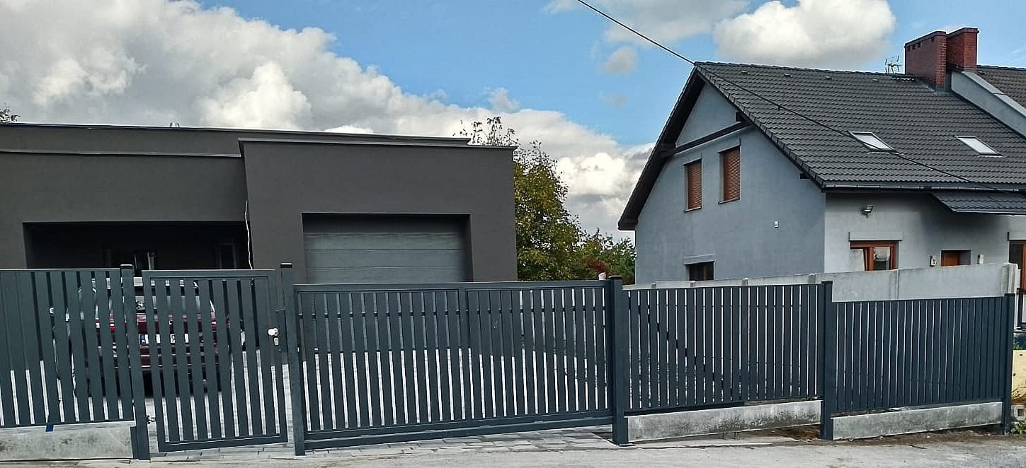 Nowoczesna, grafitowa brama wjazdowa z pionowymi szczeblami na tle domu z szarą elewacją i garażu, widoczny samochód za bramą.
