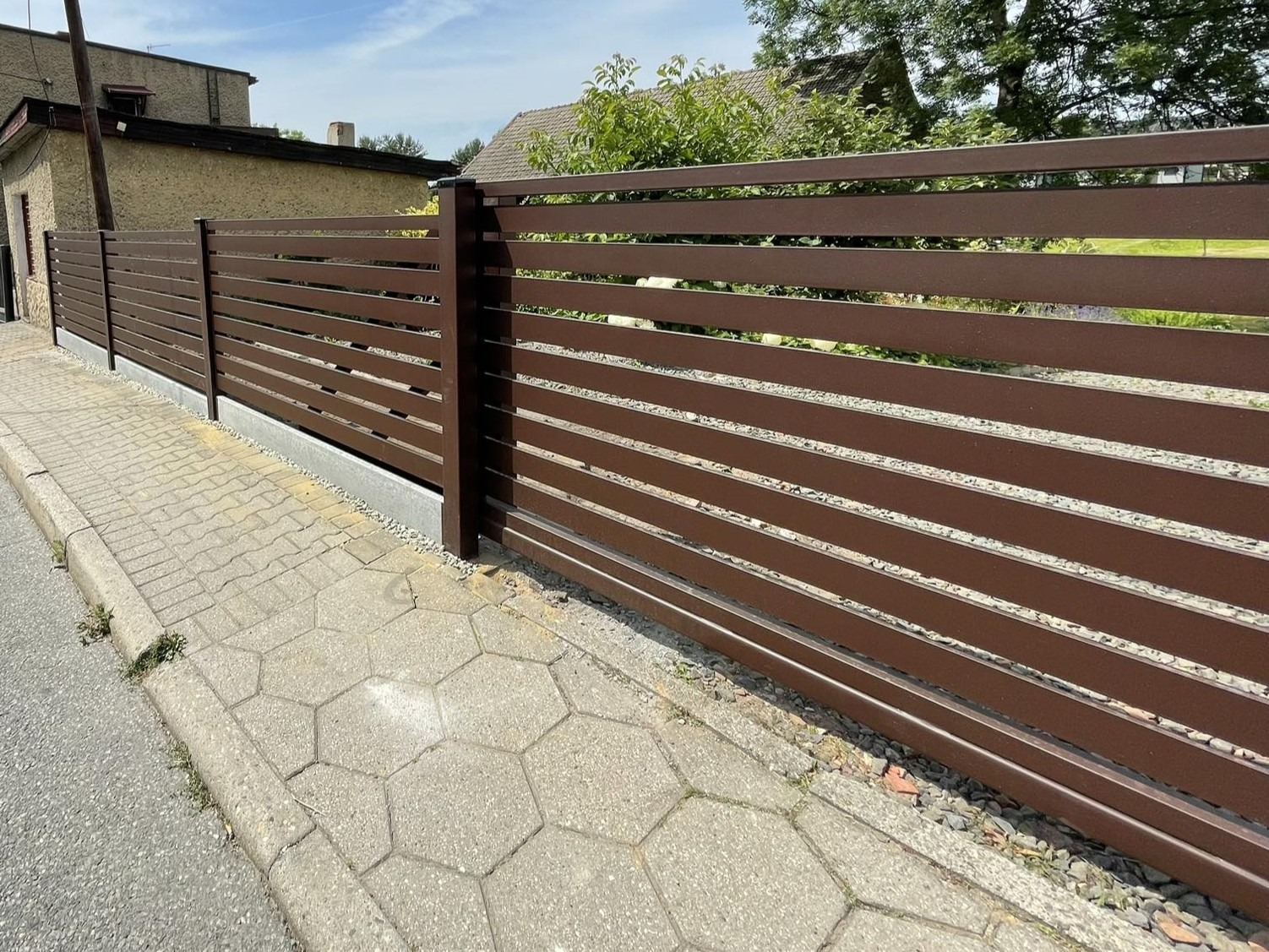 Nowoczesne, poziome ogrodzenie panelowe w kolorze brązowym, z widoczną kostką brukową i budynkiem w tle.