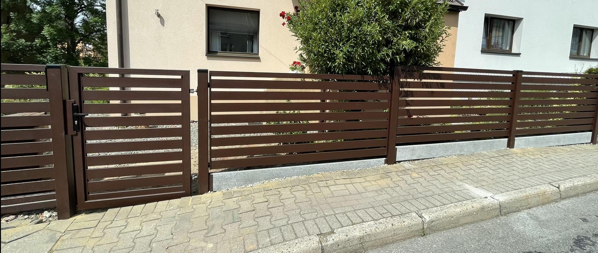 Nowoczesna, pozioma brama wjazdowa i przylegające ogrodzenie posesji w kolorze ciemnego brązu, z widoczną kostką brukową i fragmentem elewacji budynku w tle.