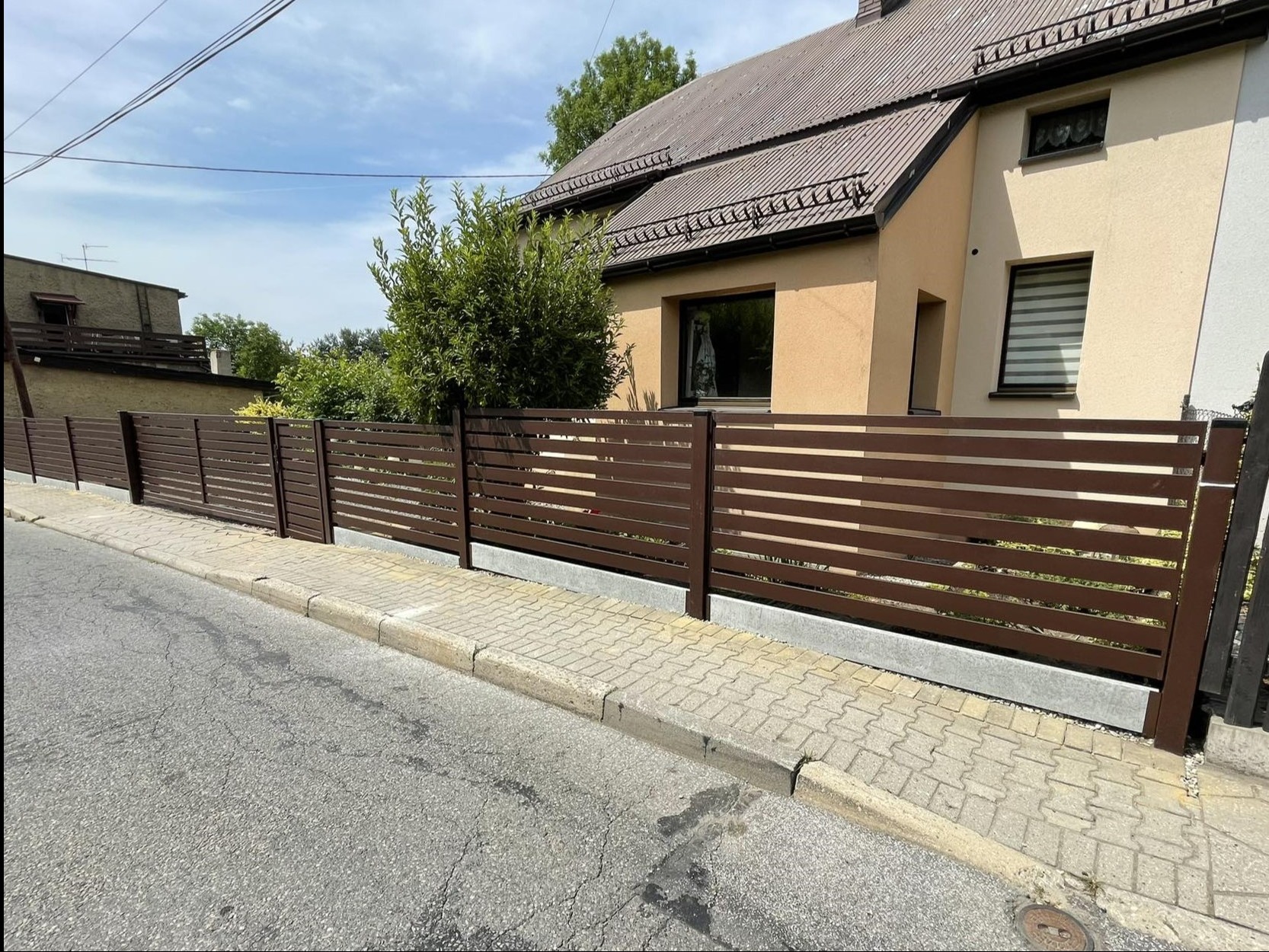 Nowoczesne ogrodzenie posesji z poziomych, brązowych profili, widoczne z perspektywy ulicy, z domem w tle.