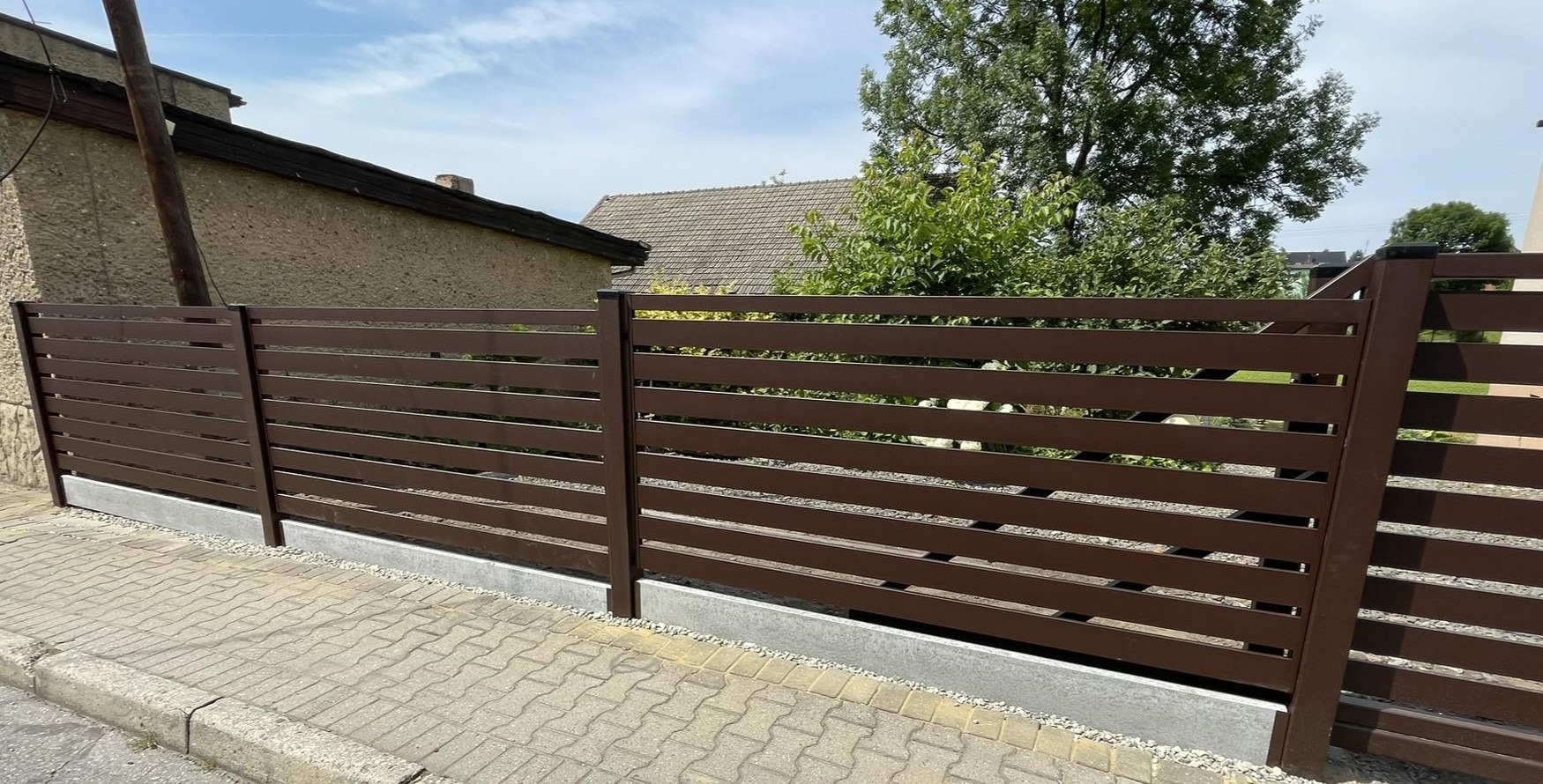 Brązowe ogrodzenie poziome z paneli na betonowej podmurówce, widok z zewnątrz na tle domów i zieleni.