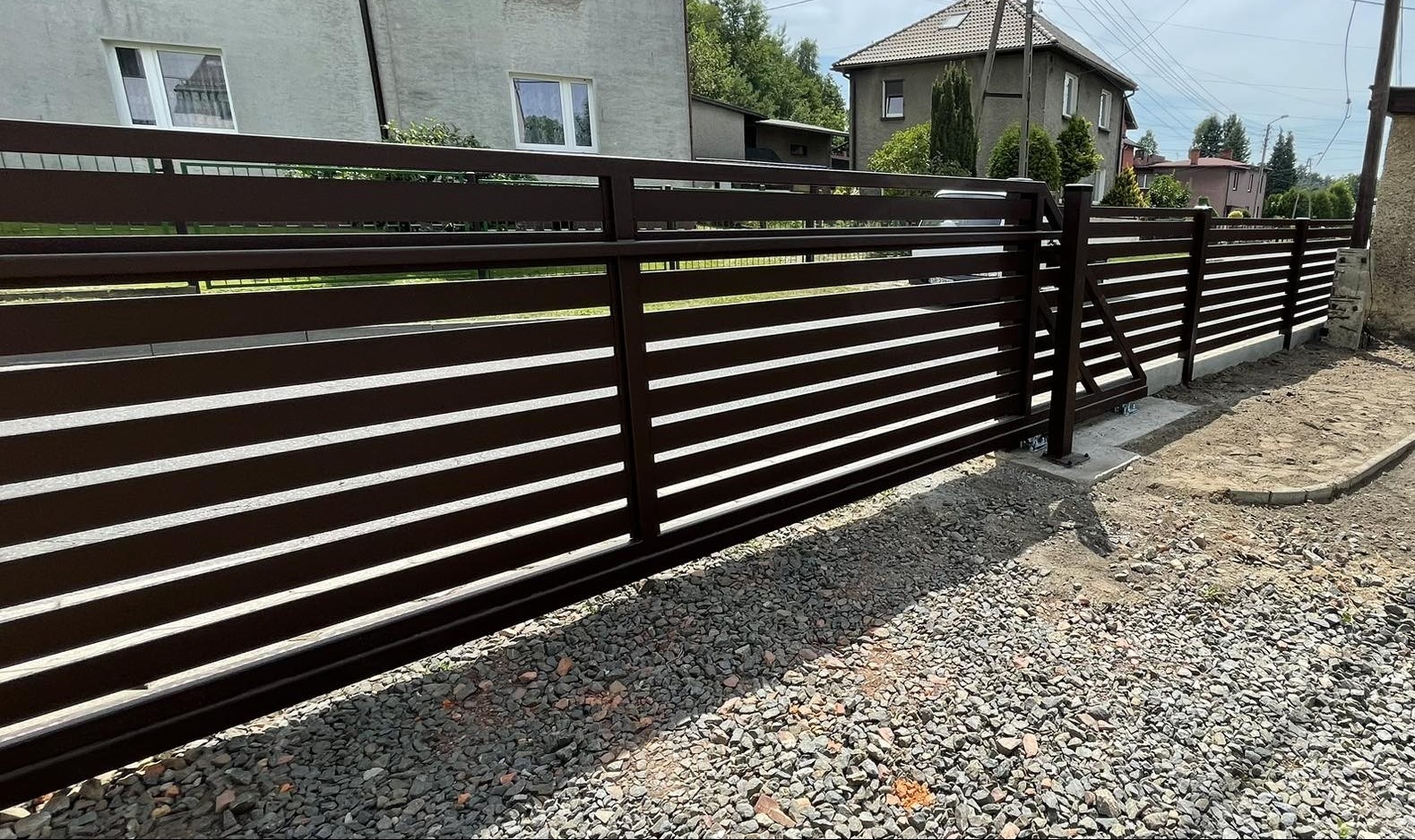 Nowoczesna, brązowa brama przesuwna z poziomymi panelami, widoczne rolki i betonowy fundament, na tle szarego domu i żwirowego podłoża.
