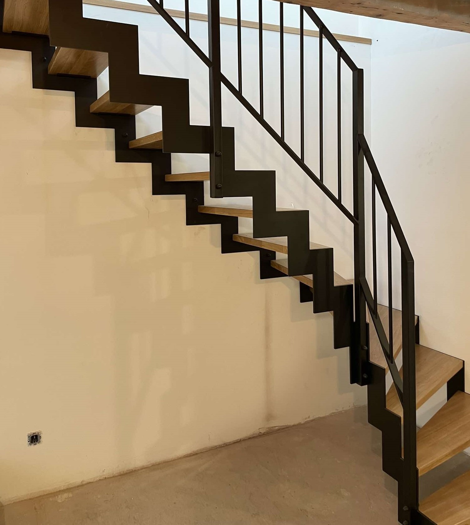 Nowoczesne schody metalowe z drewnianymi stopniami i minimalistyczną balustradą w surowym wnętrzu, widoczne gniazdko elektryczne w ścianie.