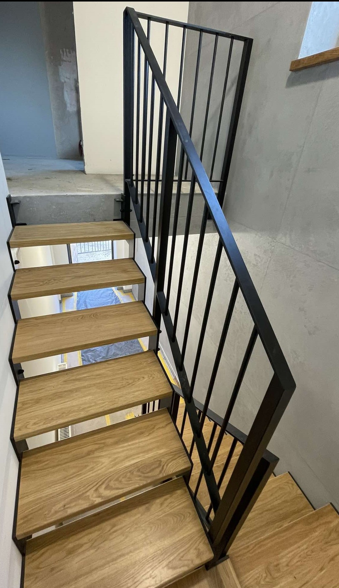 Nowoczesne schody z drewnianymi stopniami i czarną, metalową balustradą, widok z dołu, fragment podłogi zabezpieczony folią malarską.