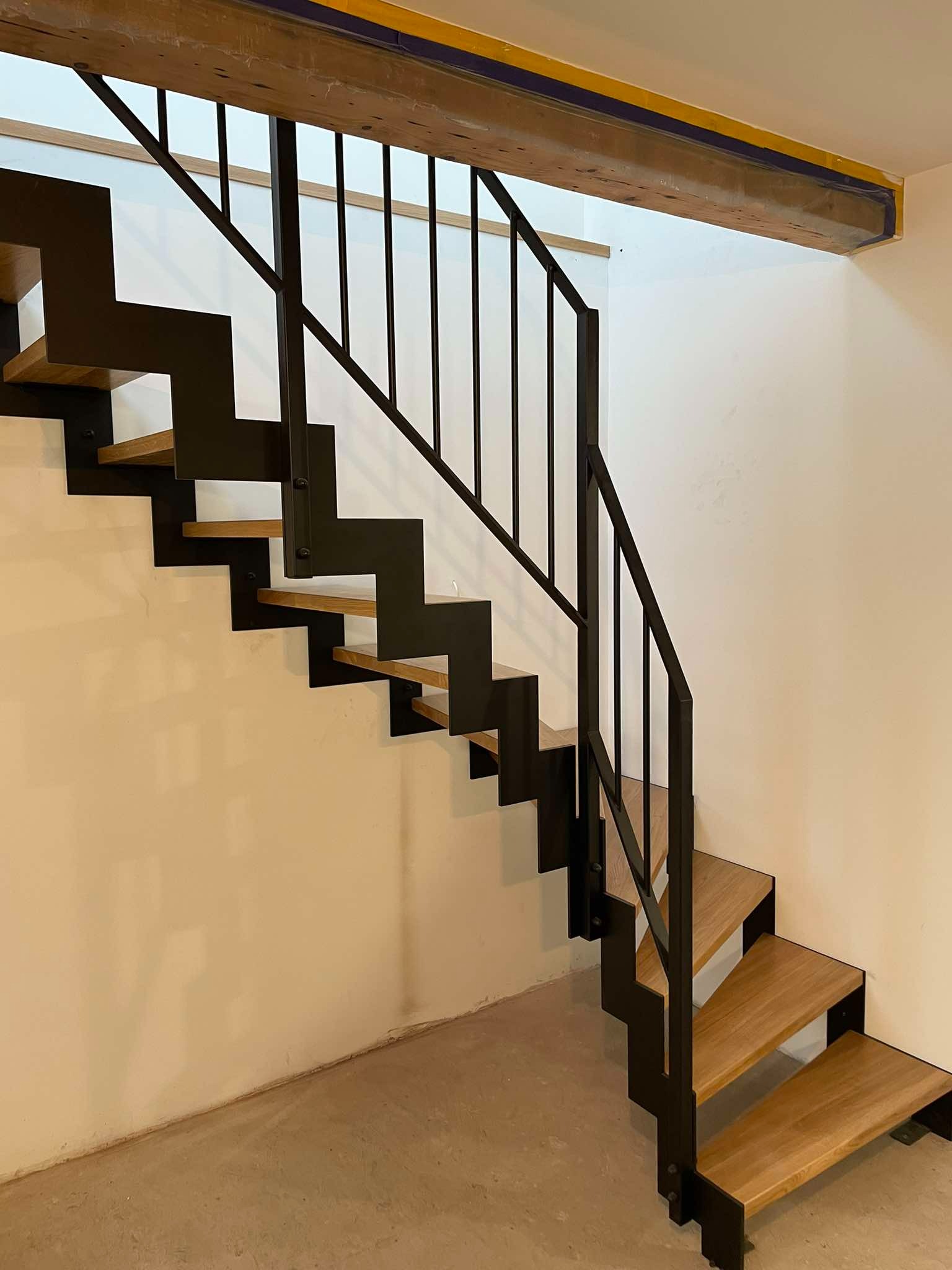 Schody metalowe z drewnianymi stopniami, czarna balustrada, widok od dołu, surowy beton na podłodze, ściany w odcieniu beżu, drewniana belka stropowa z zabezpieczeniem.
