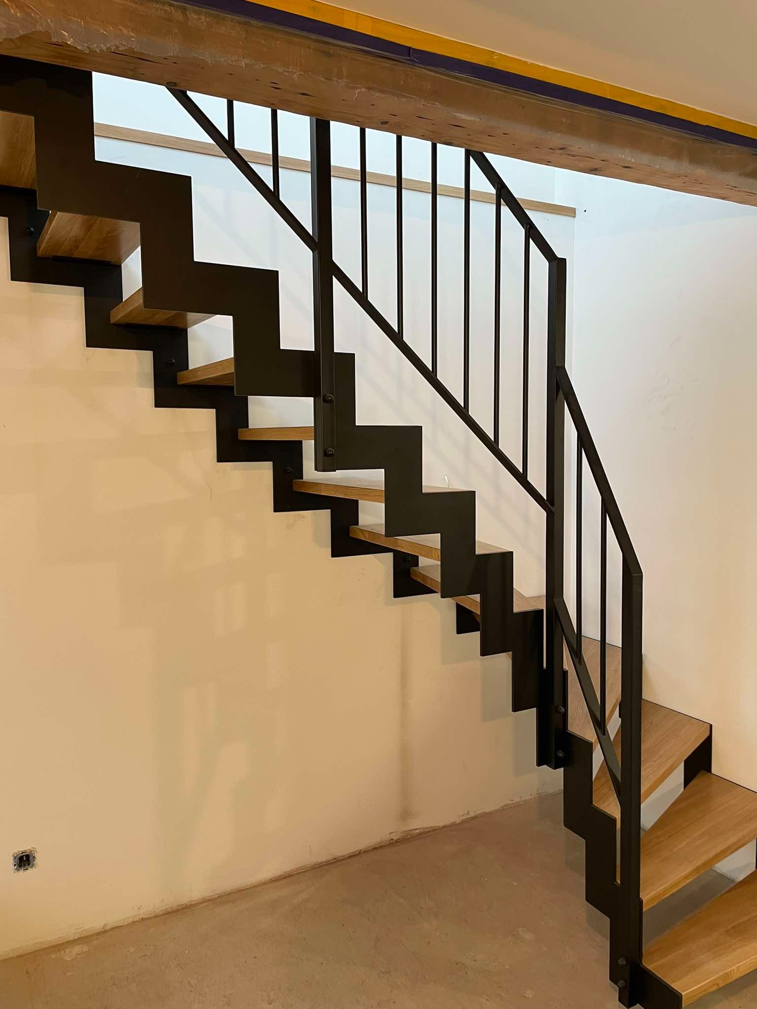 Nowoczesne schody metalowe z drewnianymi stopniami, czarne metalowe barierki, widok z dołu na jasnym tle ściany.
