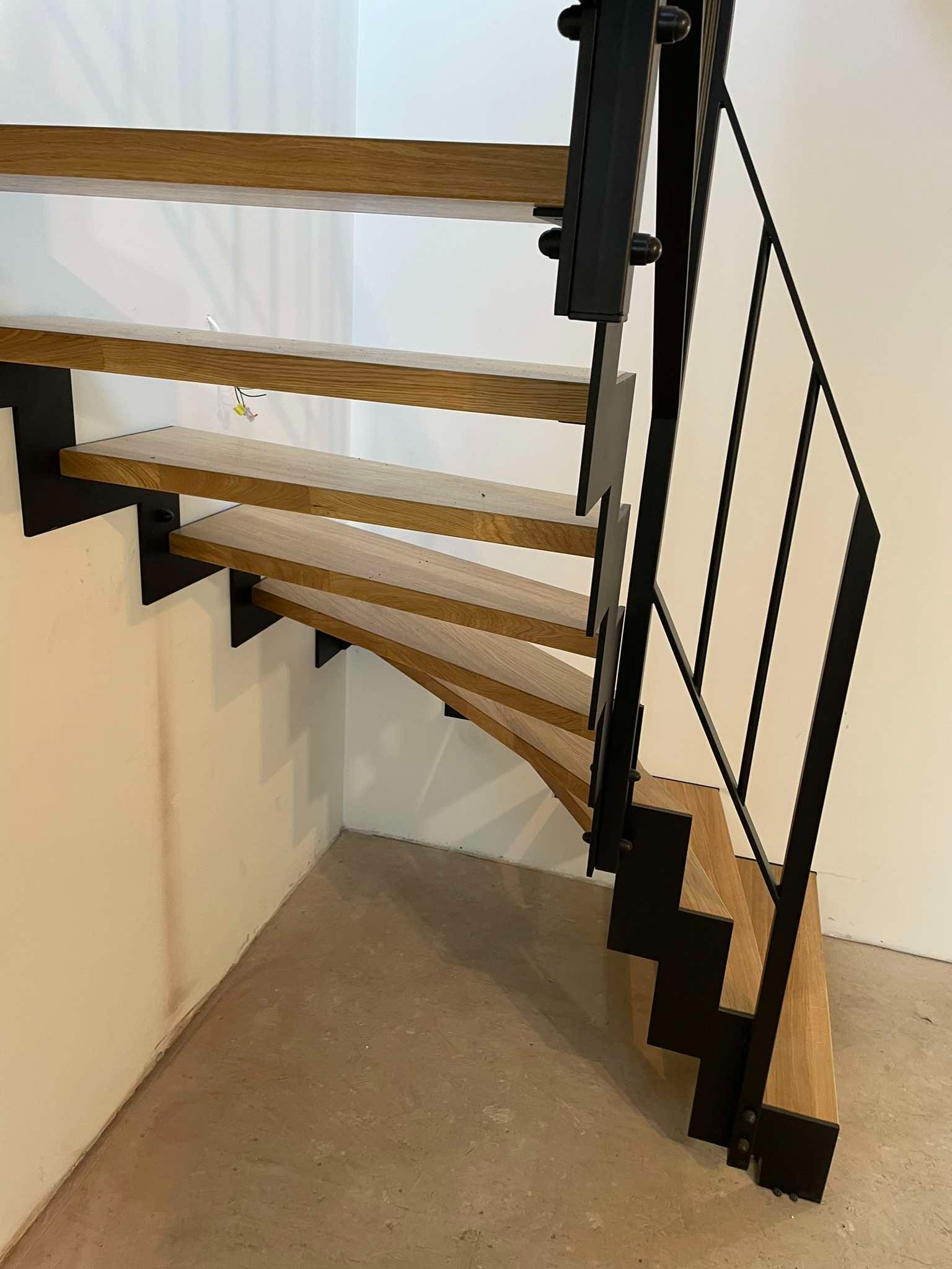 Nowoczesne schody z drewnianymi stopniami i czarną metalową konstrukcją, widok z dołu na jasnym tle ściany i betonowej podłogi.
