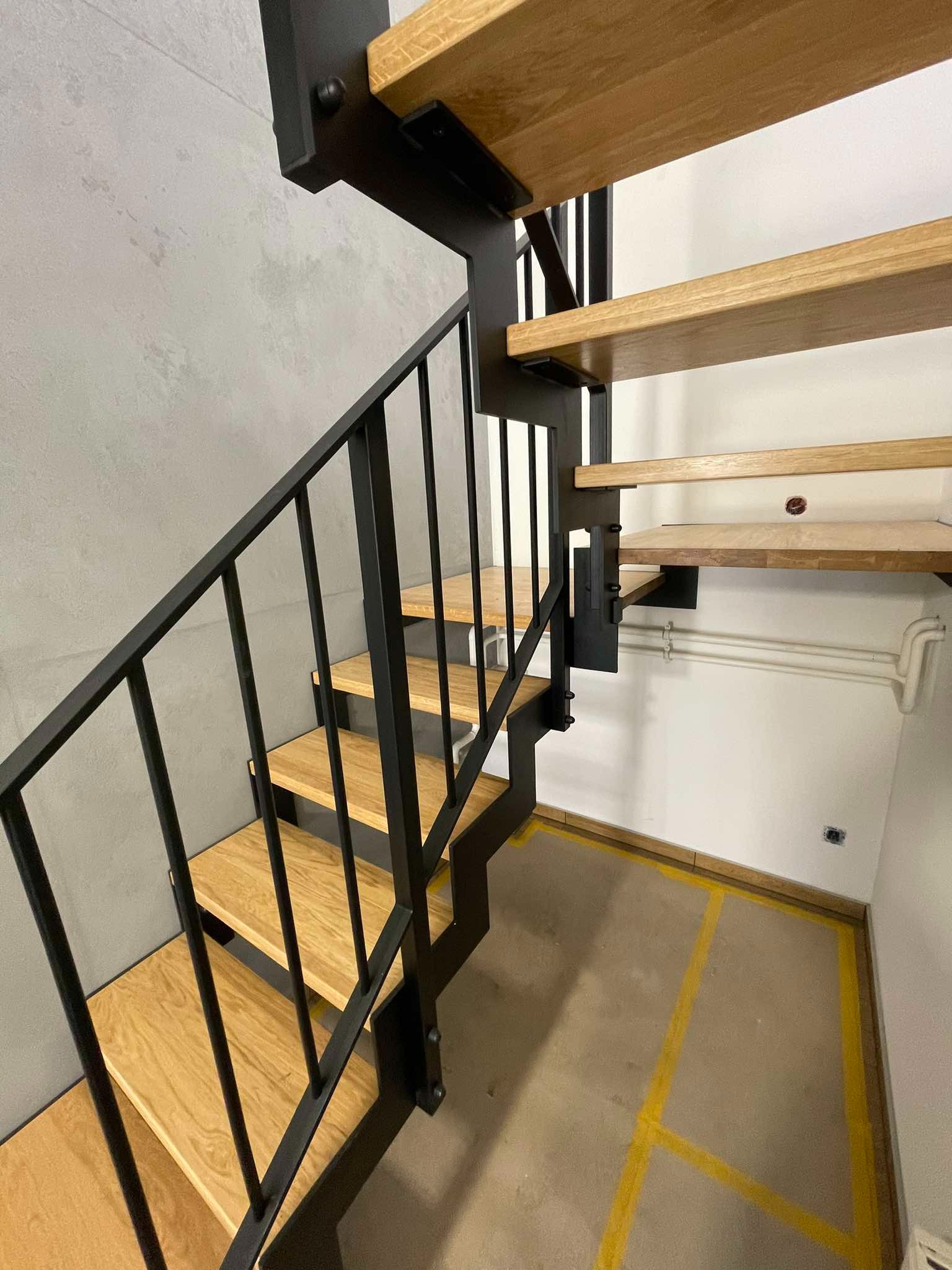 Nowoczesne schody z drewnianymi stopniami i czarną metalową konstrukcją, widok z dołu, surowy beton na ścianie, żółte linie na podłodze.