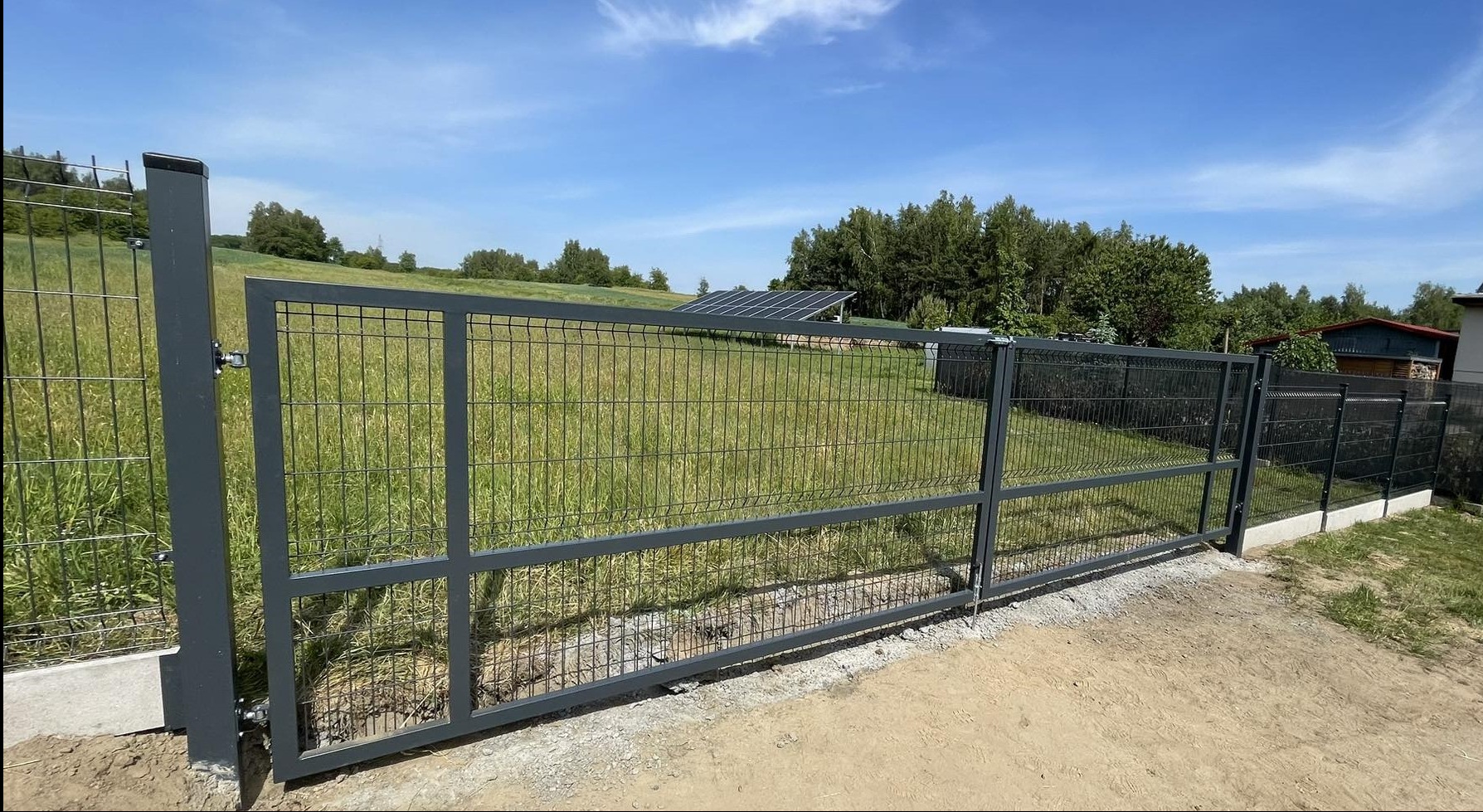 Nowoczesna, metalowa brama panelowa w kolorze grafitowym, z widocznymi spawami i zamkiem, na tle zielonego pola z panelami słonecznymi w oddali, brama częściowo otwarta.